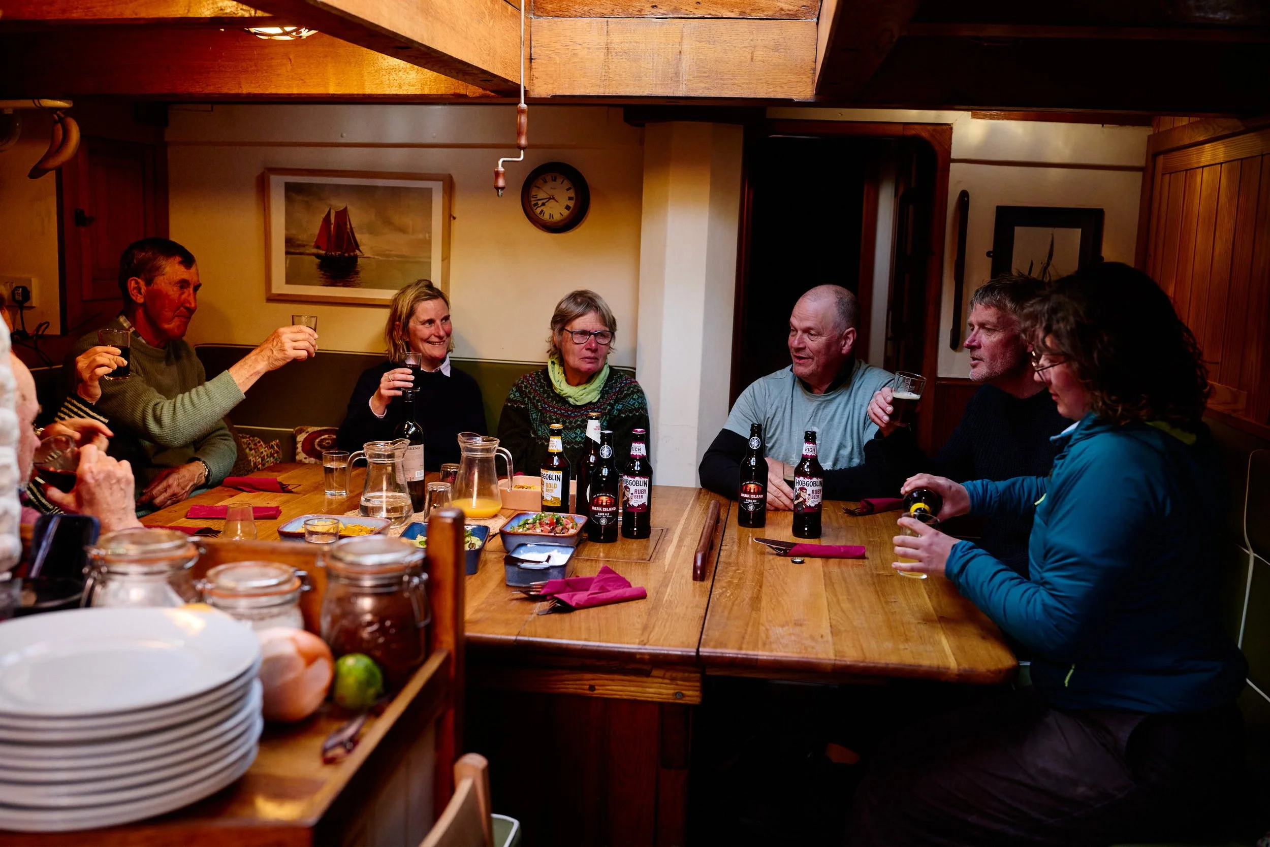 A group of people sitting around a large wooden dining table enjoying drinks and conversation in a cozy, warmly lit room. The table has various dishes and drinks, and the background features framed paintings, a clock, and wooden wall paneling.