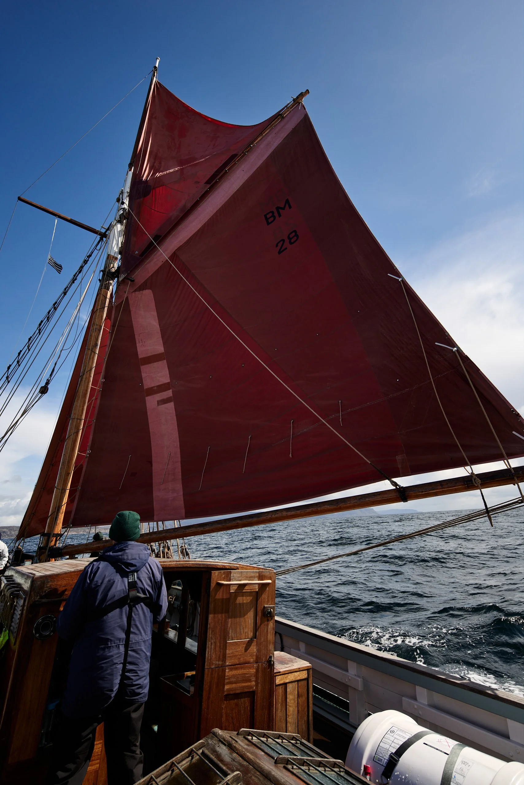 Sailboat with red sails on open water with a crew member wearing a green hat and navy jacket looking forward.