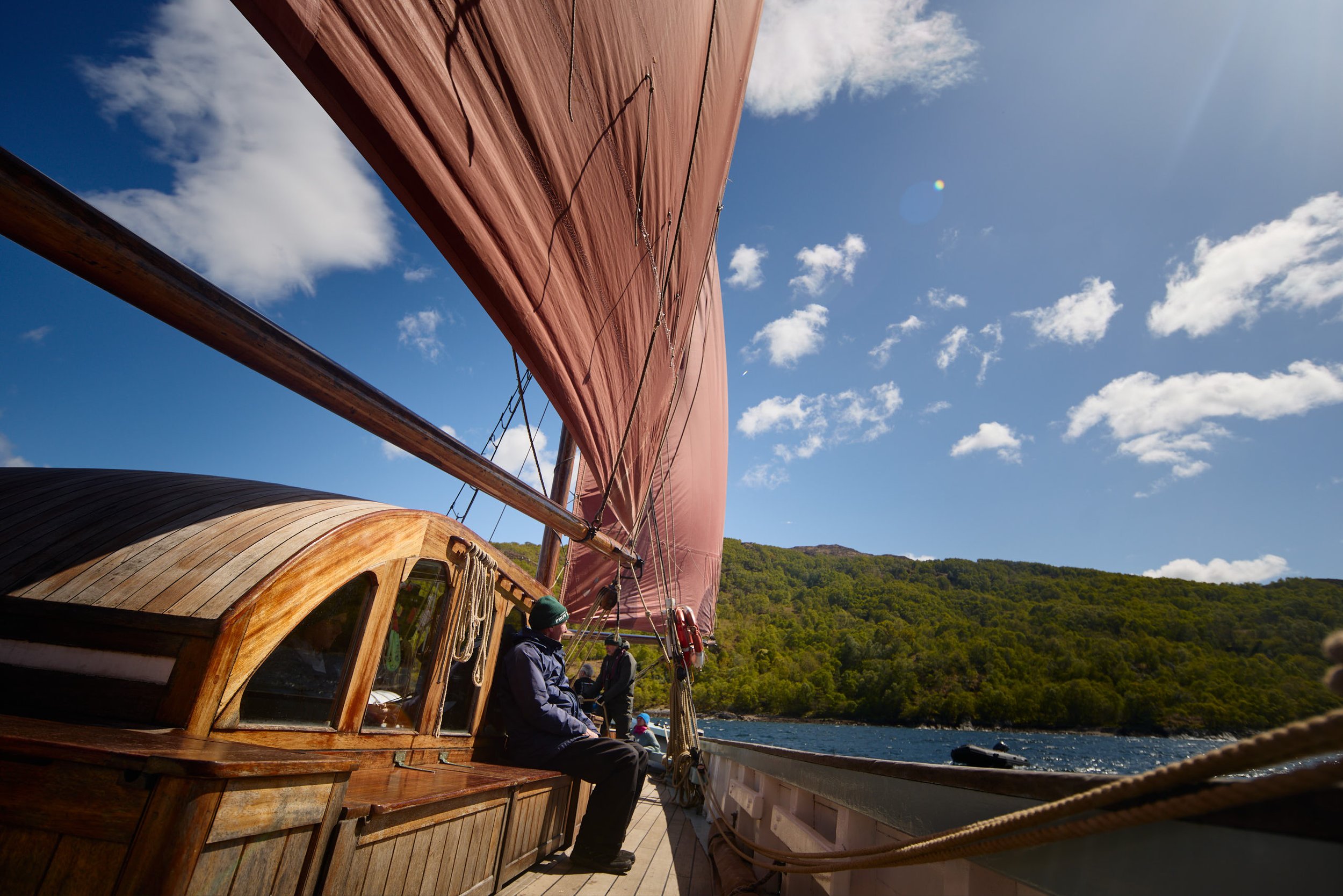 View from the deck of a historic wooden sailing ship with a large red sail, overlooking a body of water and lush green hills under a blue sky with scattered white clouds.