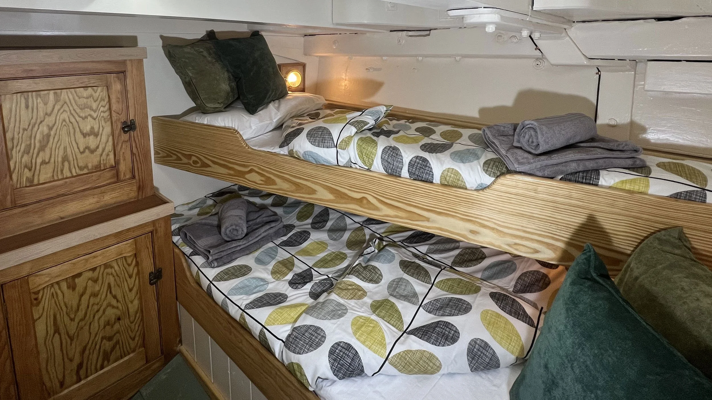 A cozy bunk bed with two beds, featuring patterned bedding with multicolored circles. The top bunk has two pillows, and a gray towel is rolled up on the right side of the bed. Next to the bunk bed, there is a wooden cabinet, and a small wall-mounted light is turned on above the top bed.