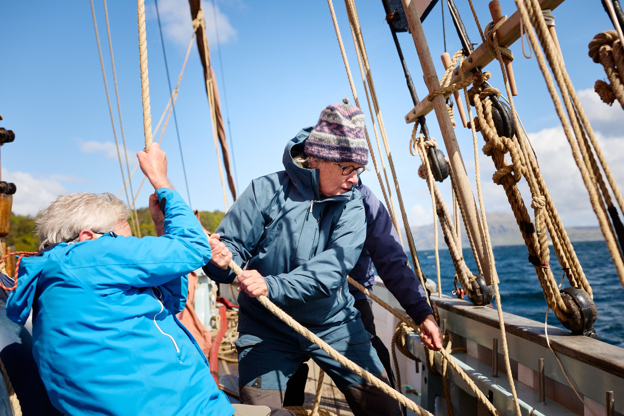 People working together on a sailing ship, holding ropes, with a background of blue sky and water.