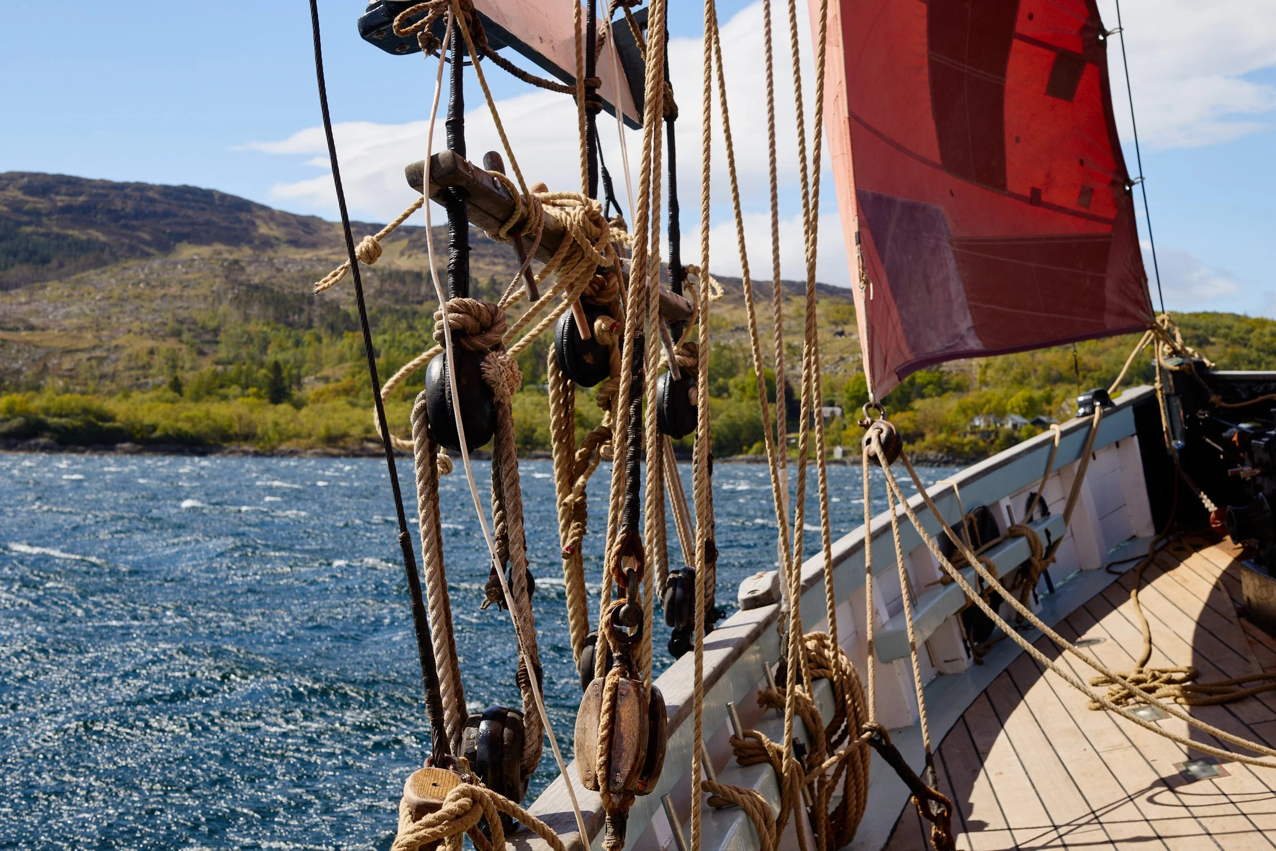 Close-up of ropes and pulleys on a wooden sailing ship with a red sail, sailing on choppy water near a rocky, forested coastline under a partly cloudy sky.