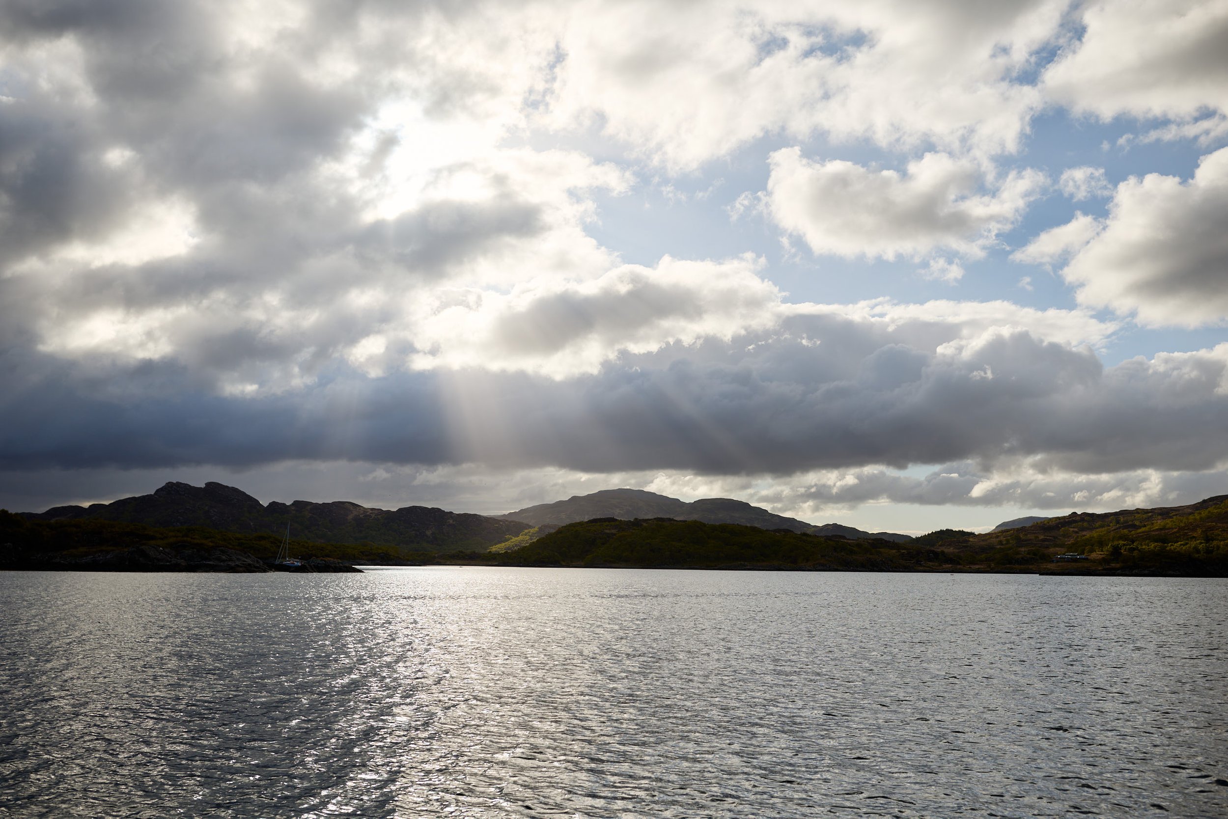 A scenic landscape featuring a body of water in the foreground, rolling hills and mountains in the background, and a partly cloudy sky with sunlight breaking through the clouds.