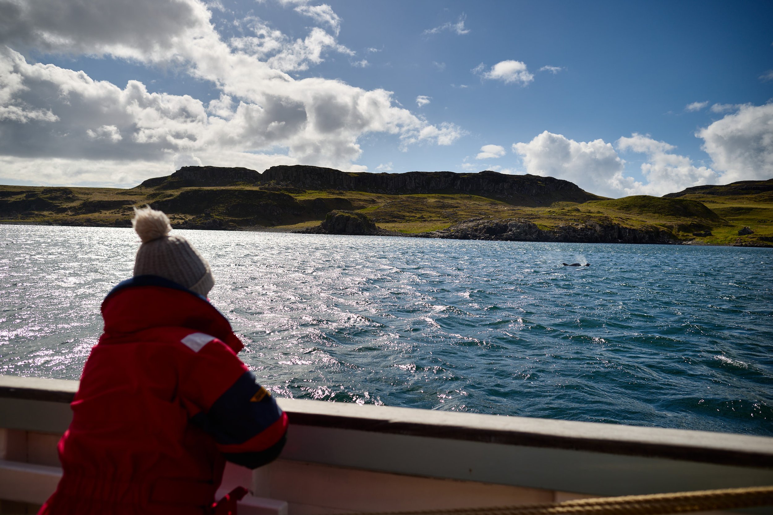 A person in a red jacket and beige beanie looking over a body of water, with grassy hills and rocky cliffs under a partly cloudy sky.