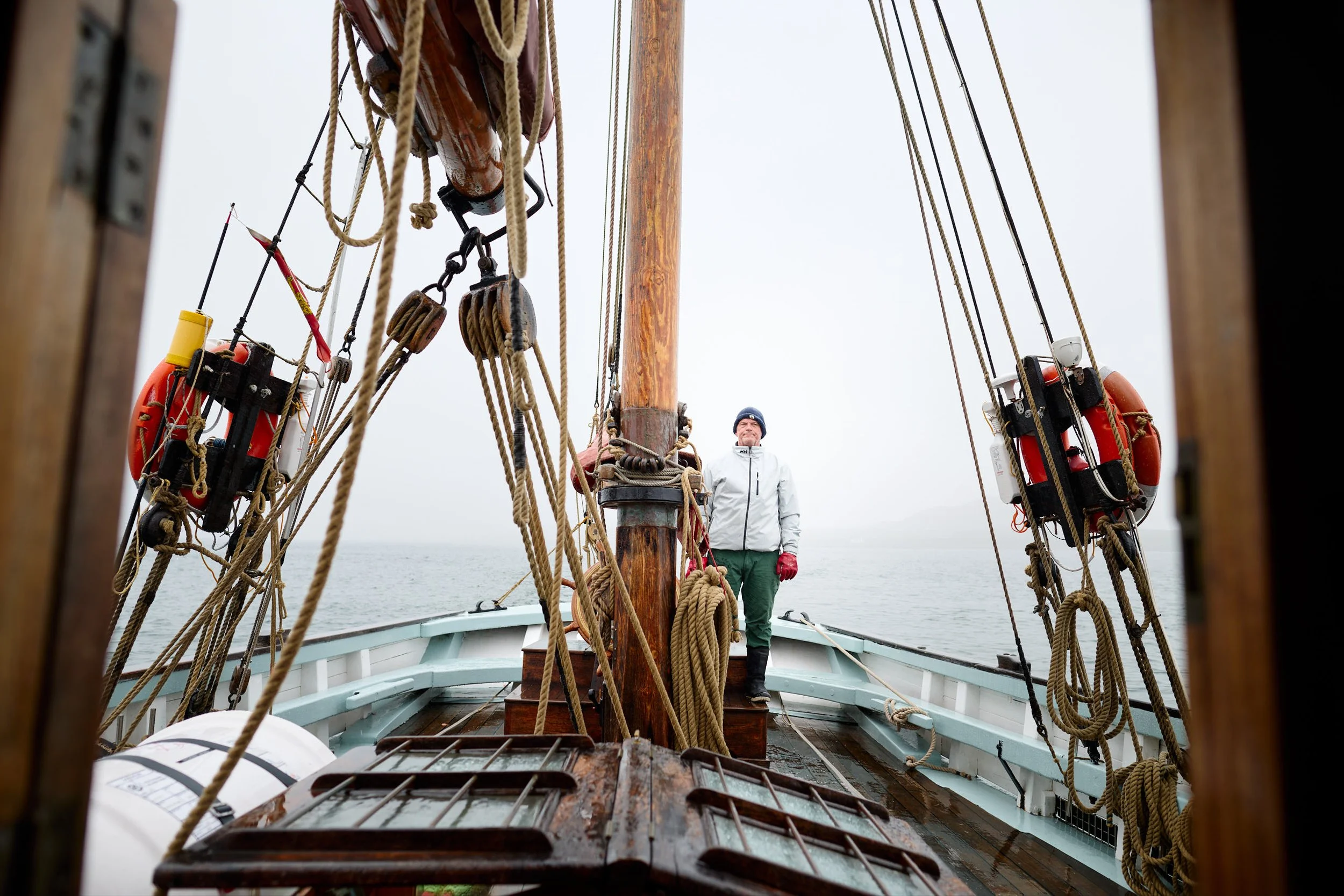 A person standing on the deck of a wooden sailing ship with numerous ropes, pulleys, and life preservers, looking out at the water under an overcast sky.