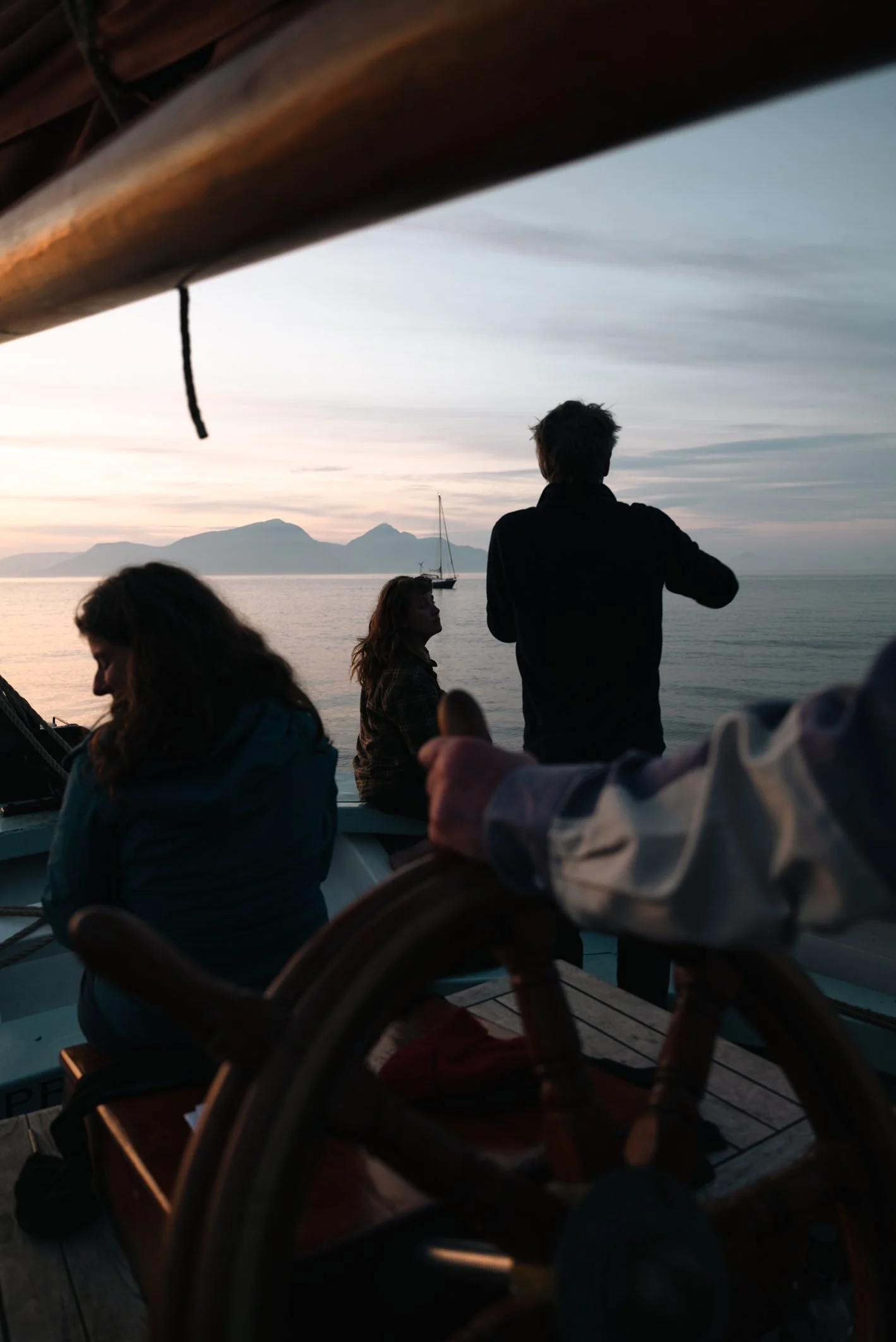 People sitting on a boat during sunset, with mountains and water in the background, and a sailboat on the water.