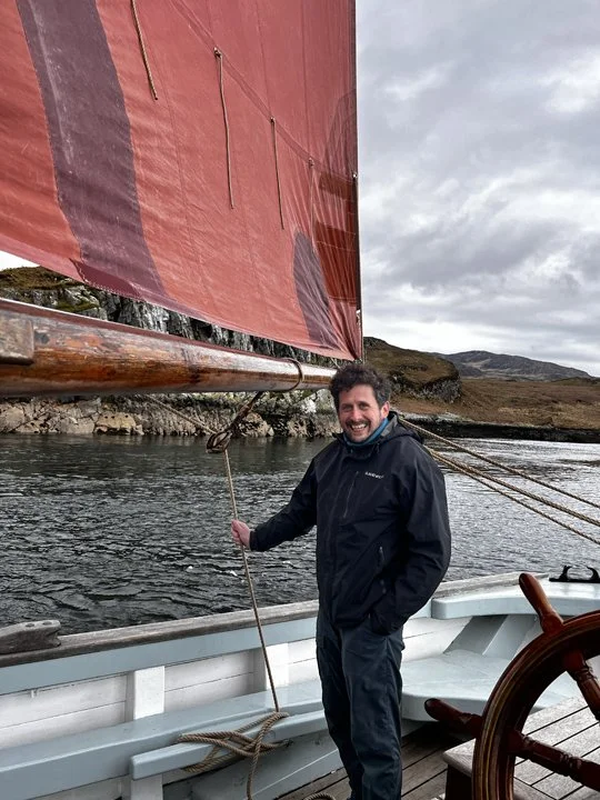 A smiling man standing on a boat holding a rope with a large red sail in the background, with water and hills under cloudy skies.