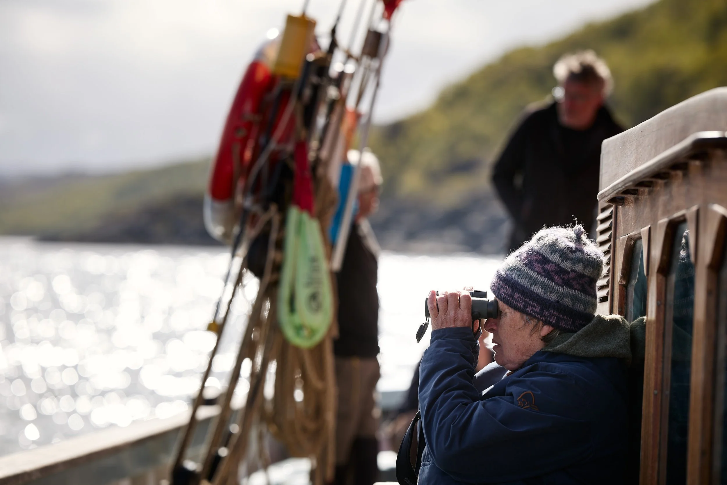 A person wearing a striped beanie and blue jacket looking through binoculars on a boat, with another person in the background near a fishing rod.