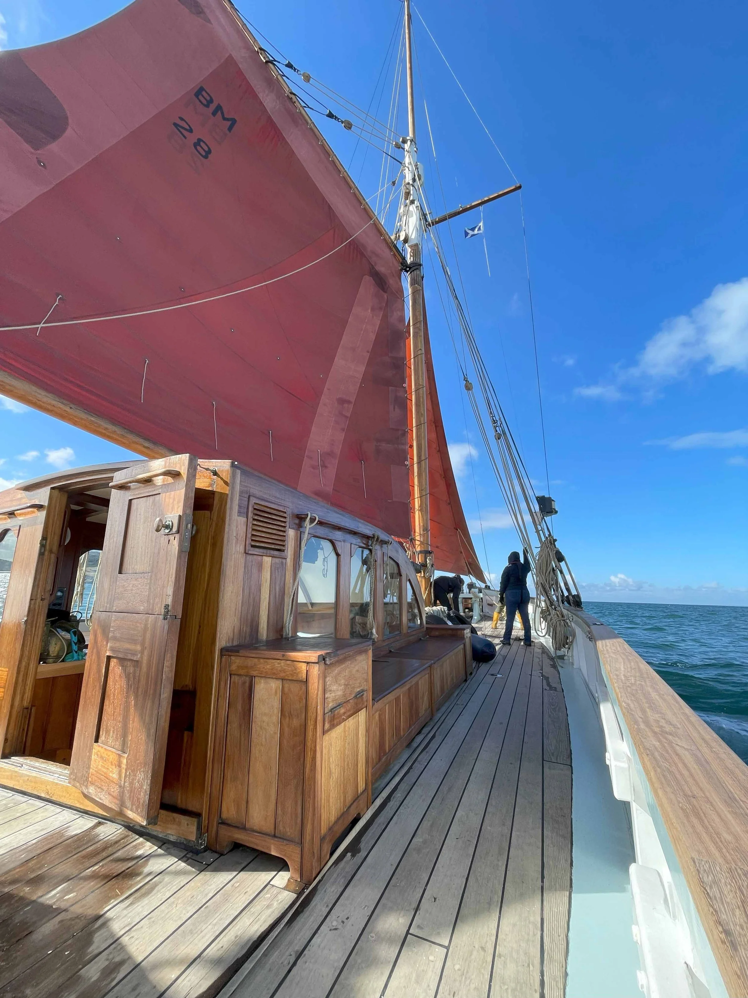 A wooden sailing ship deck with a crimson sail, a wooden cabin, and several people standing near the railing, with a blue sky and ocean in the background.