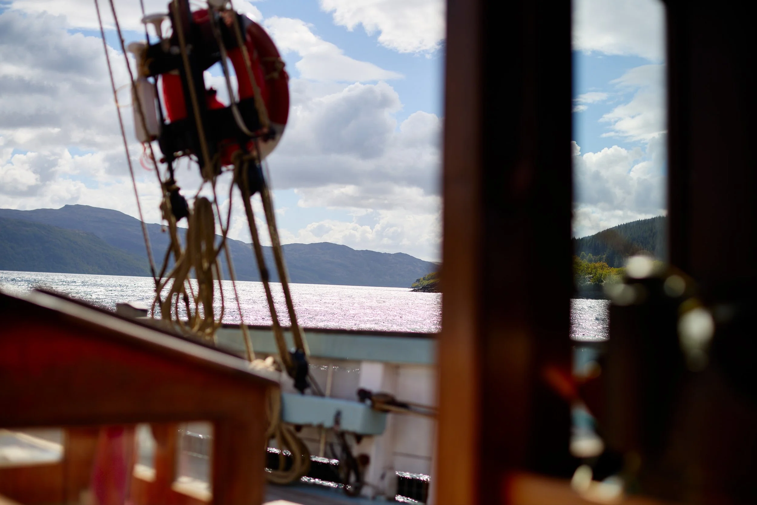 A view of a lake with mountains and a partly cloudy sky, seen through the window of a boat, with a lifebuoy hanging on the boat's side in the foreground.