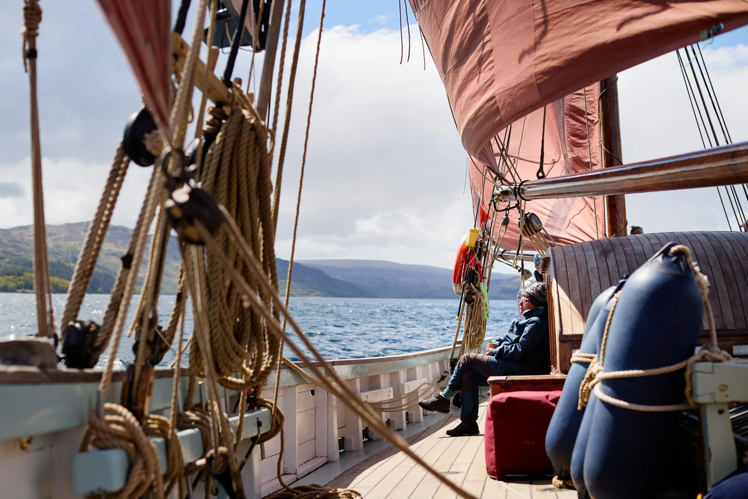 A person sitting on a boat with a wooden deck, surrounded by ropes and rigging, with mountains and water in the background under a partly cloudy sky.