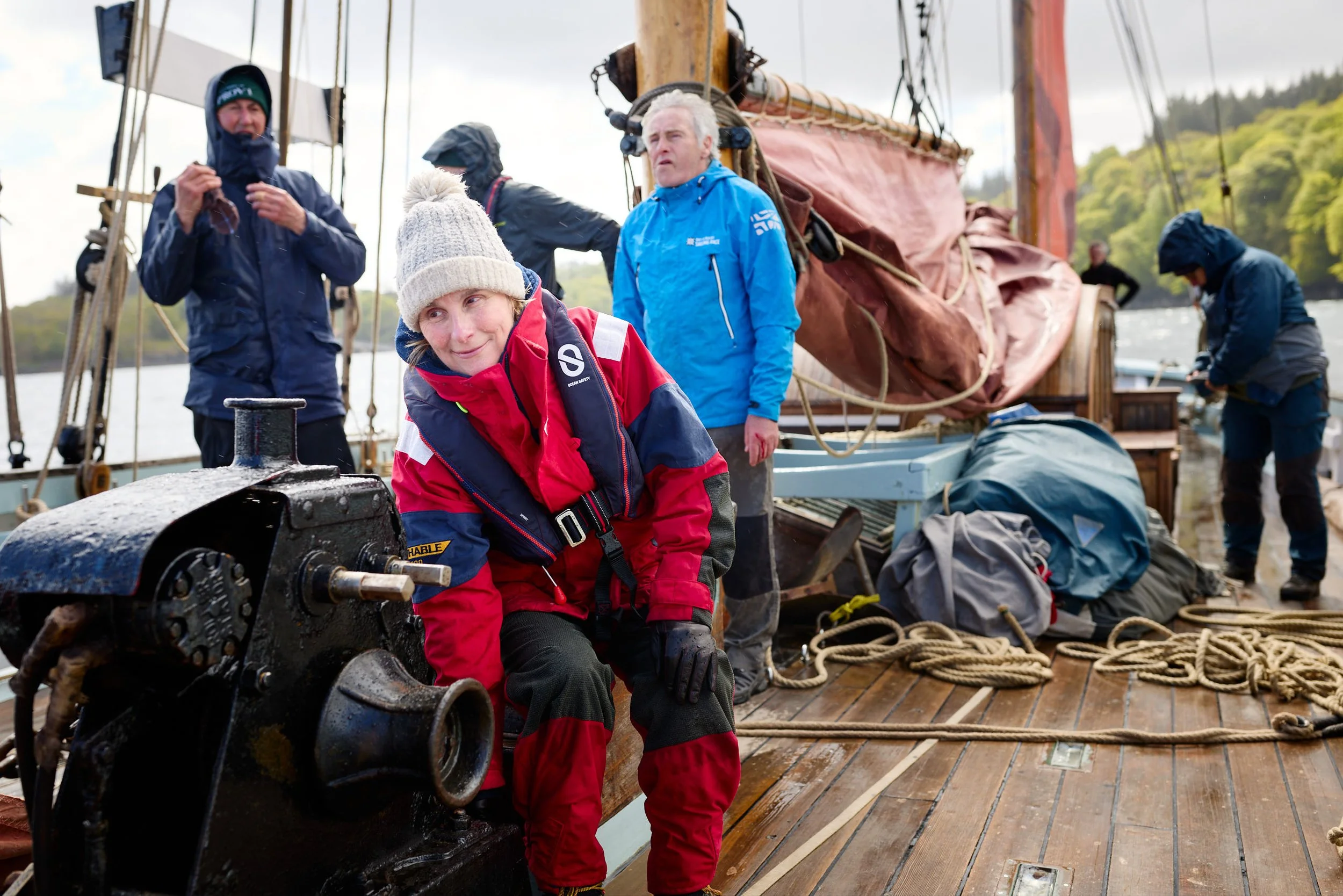 A group of people on a boat dressed in rain jackets and gear, with some working and others relaxing, on a cloudy day.
