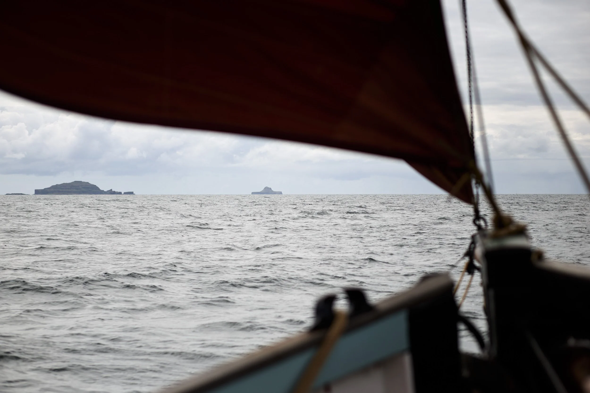 View of the ocean with distant islands seen through a boat's sail and rigging, partially covered by the boat's dark canopy.