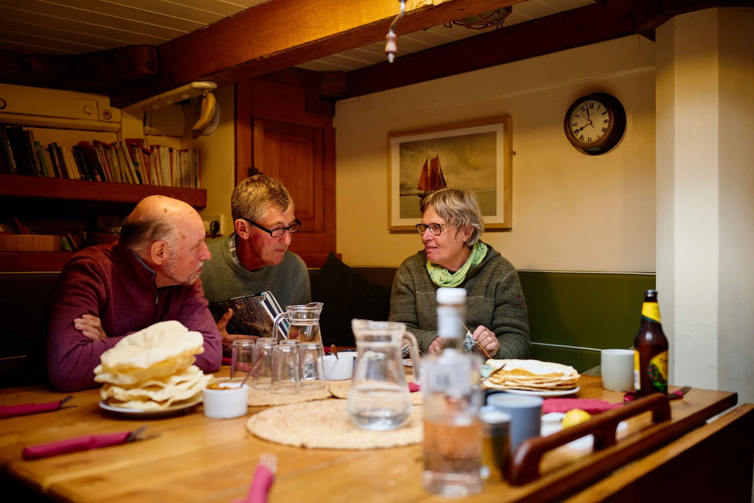 Three older adults sitting at a long wooden dining table having a conversation. The table has plates of flatbread, glasses, and bottles. The background has a wall with a beach-themed painting and a clock. The room has wood accents and a shelf with books.