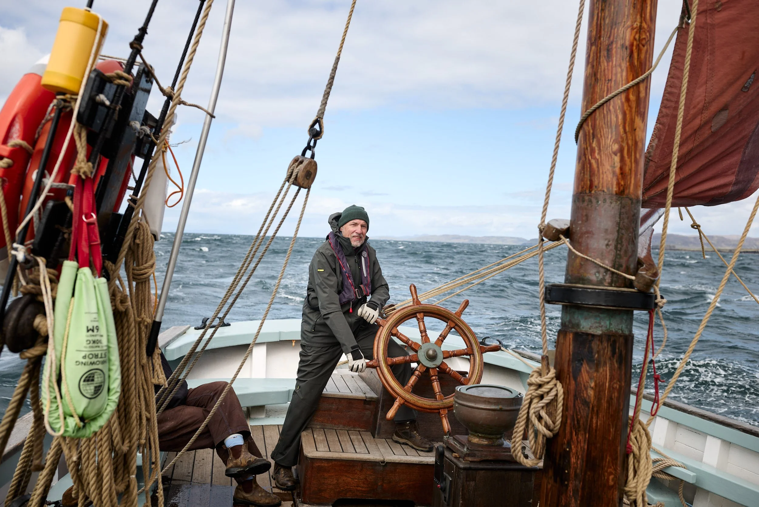 A man in a green beanie and gray jacket steering a sailboat with a wooden wheel, surrounded by ropes and nautical equipment, on open water under a partly cloudy sky.