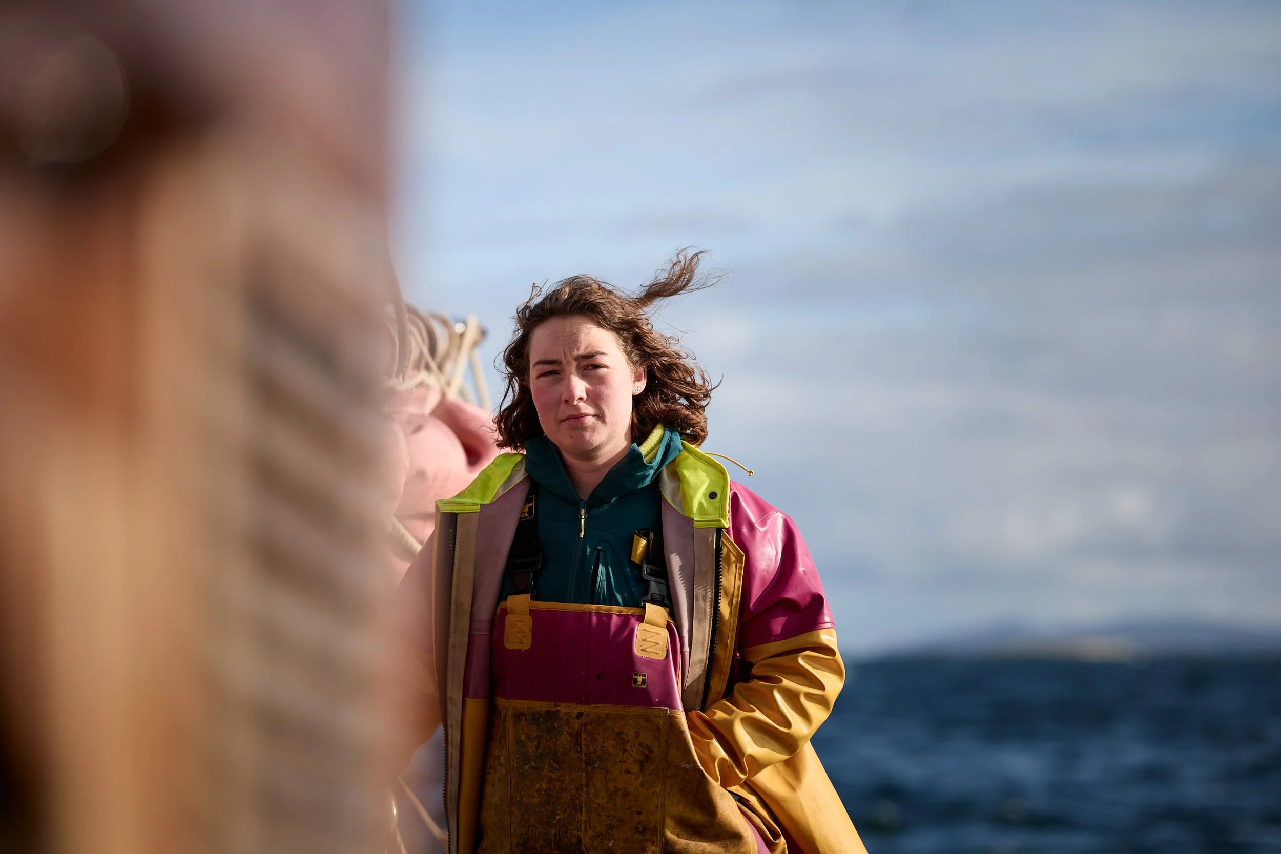 A woman with wavy brown hair wearing a colorful waterproof jacket and a life vest, standing outdoors with the ocean and a cloudy sky in the background.