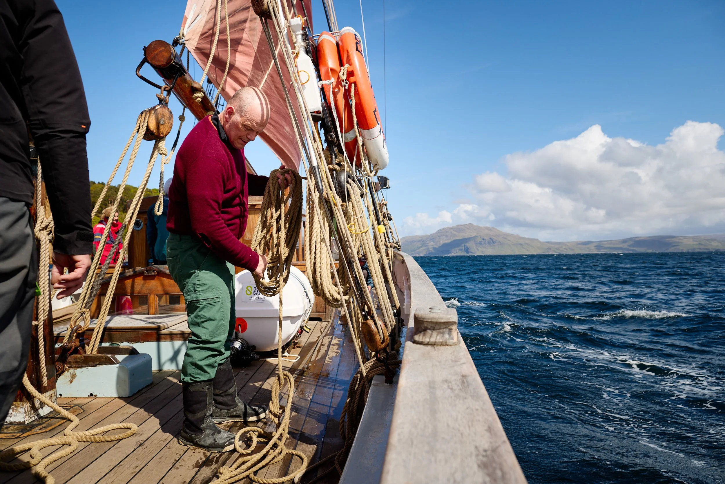 Older man on a wooden boat, handling ropes, with a backdrop of open water and distant hills under a partly cloudy sky.