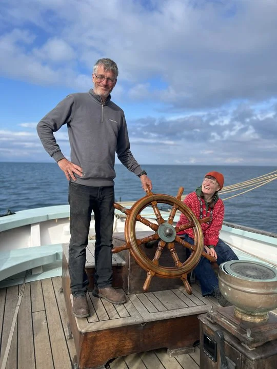 A man and a woman on a boat. The man is standing and smiling, holding a boat's ship wheel. The woman is sitting, wearing glasses and a red cap, and laughing. The ocean and sky are visible in the background.