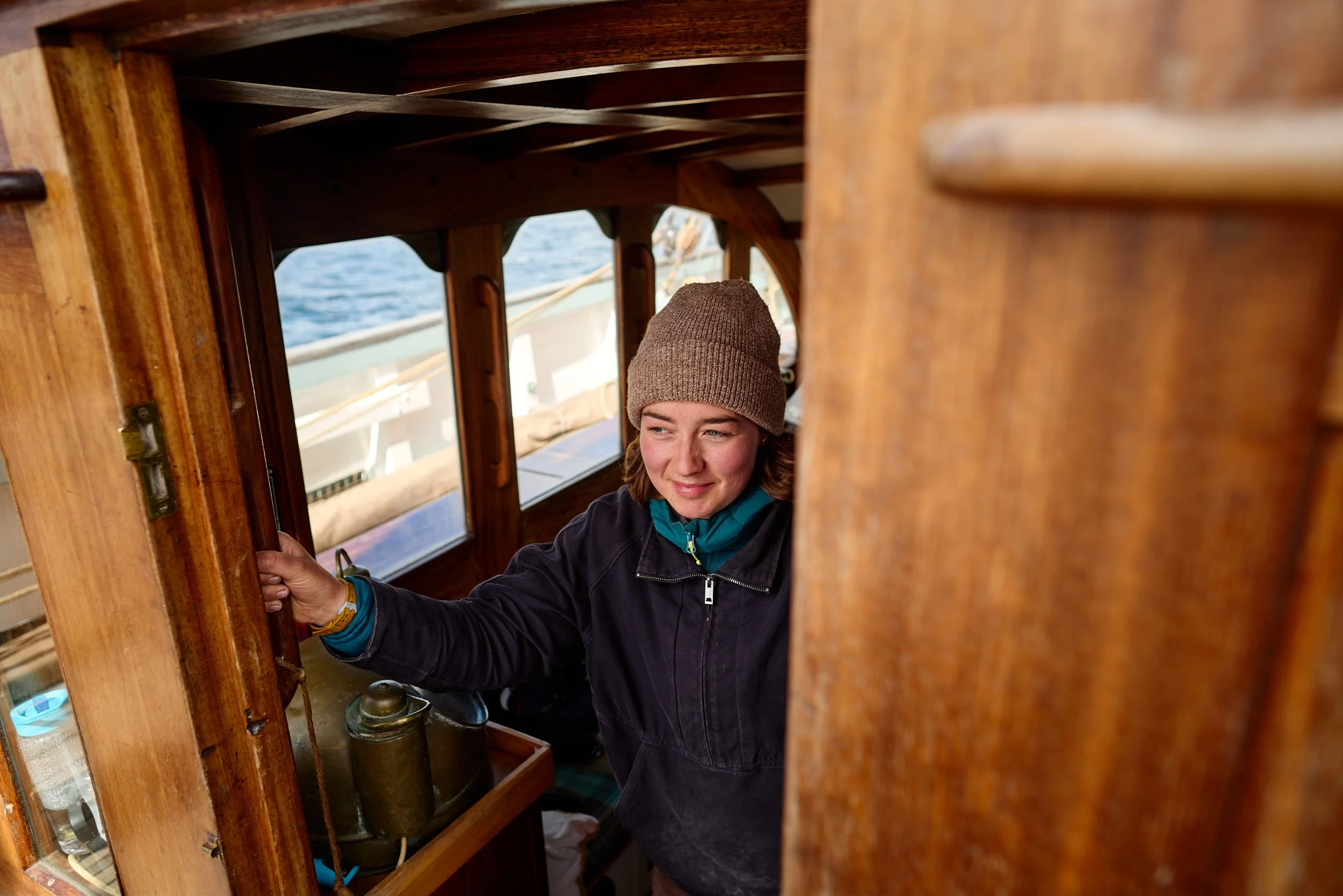 A woman wearing a brown beanie and black jacket looking out of a small boat's wooden window inside cabin, with water visible outside.