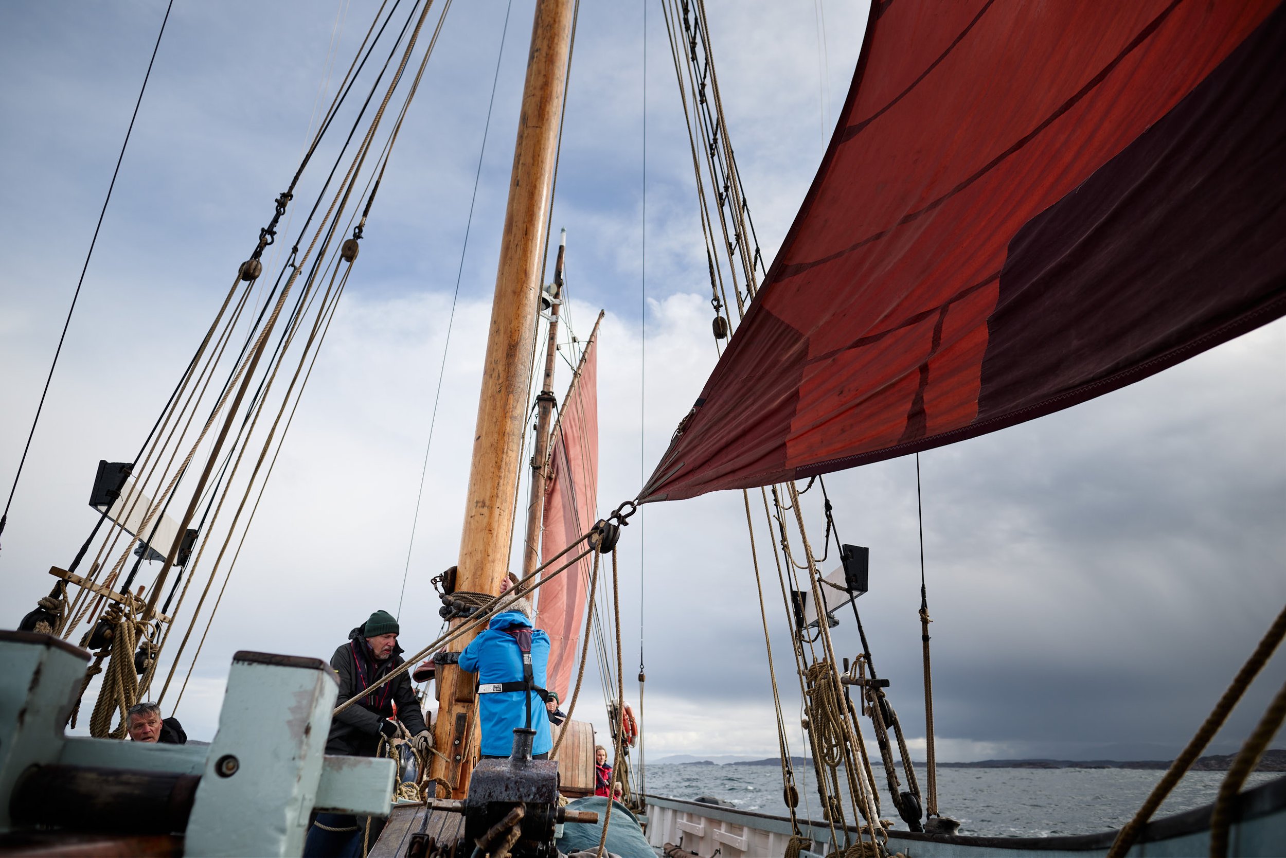 People working on a sailing ship with wood masts and orange sails, on a cloudy day with water and distant land in the background.