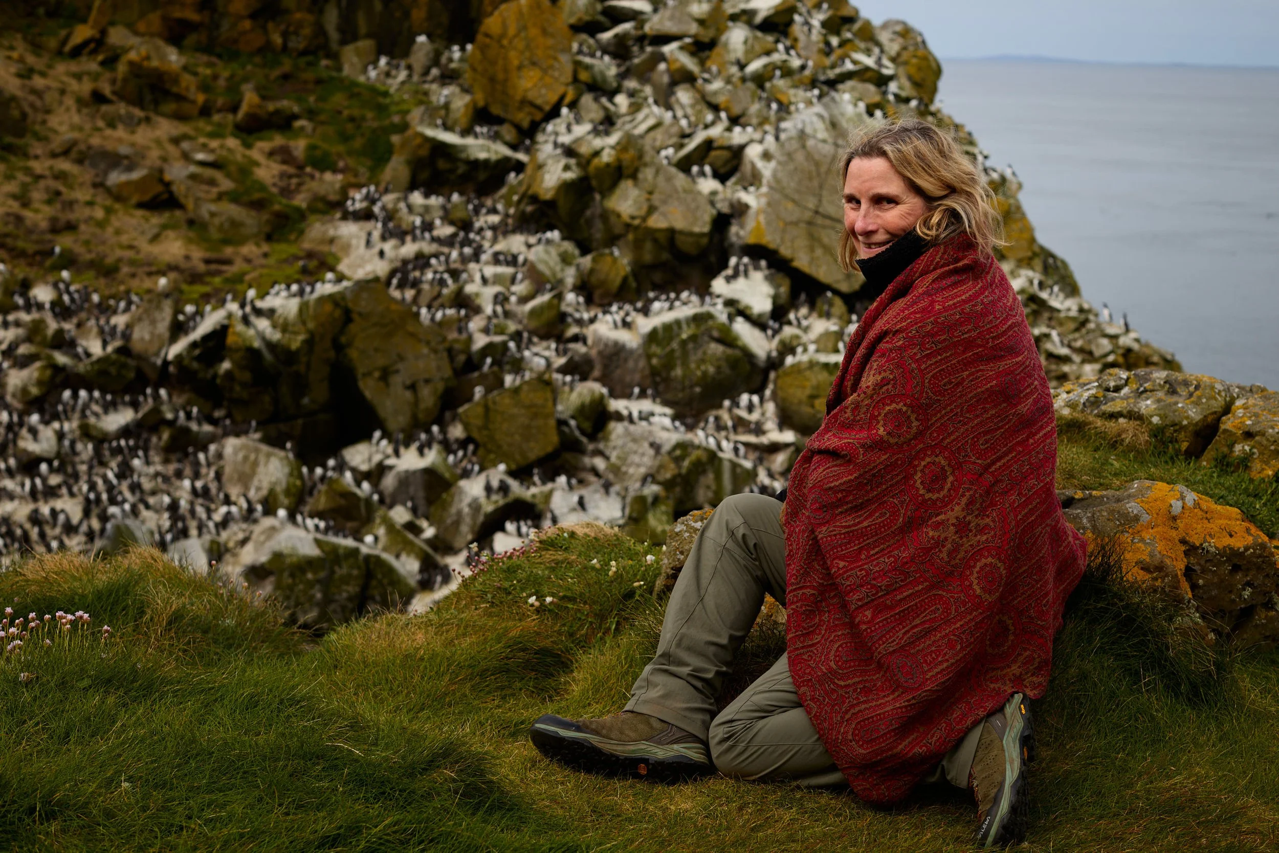 Woman seated on grassy hillside near rocky cliffs with a large blanket draped over her shoulders, surrounded by wildflowers, with a cloudy sky and ocean in the background.