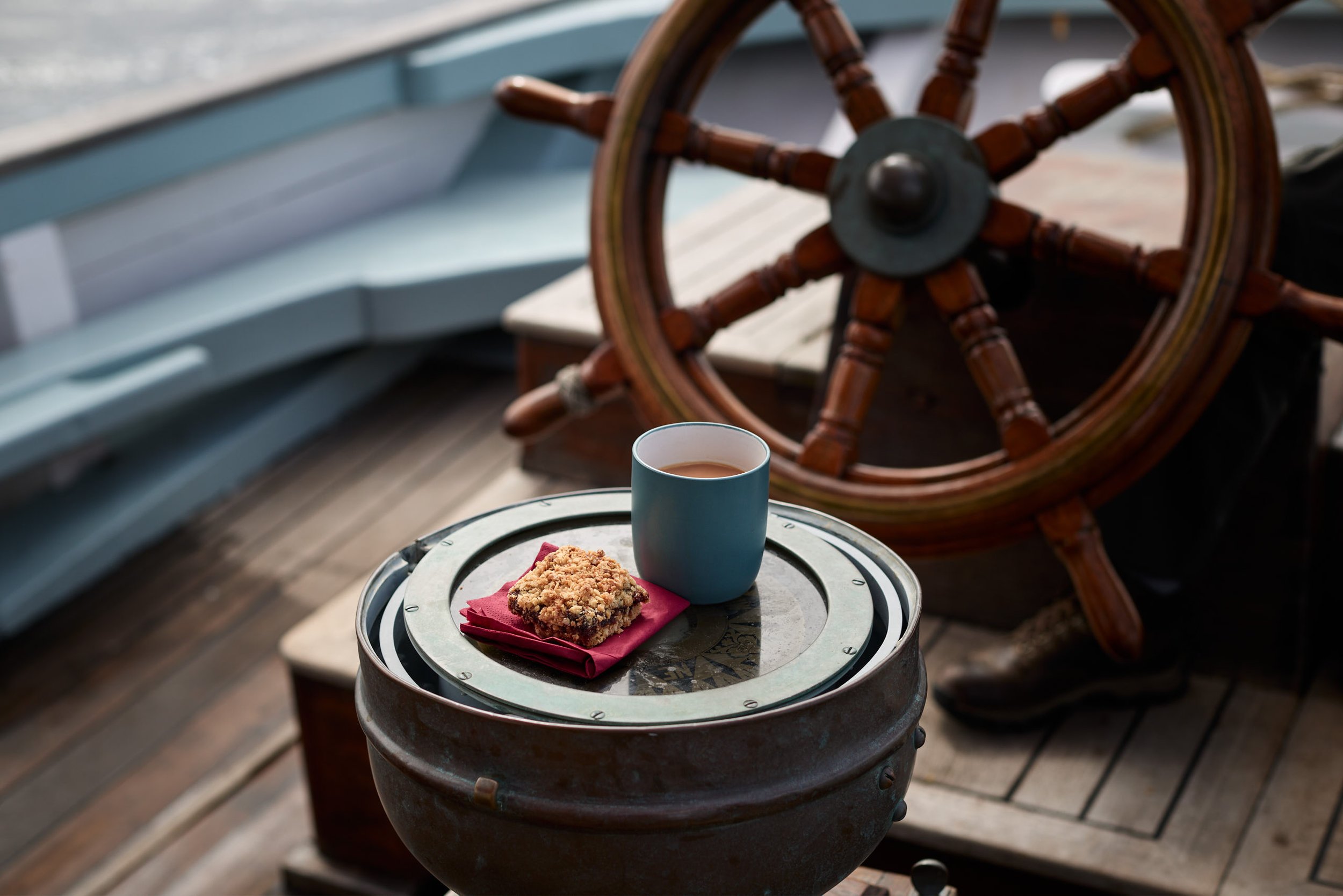 Compass with a coffee cup, a cookie, and a red napkin, set on a round metal container on a wooden deck, with a ship's wheel in the background.