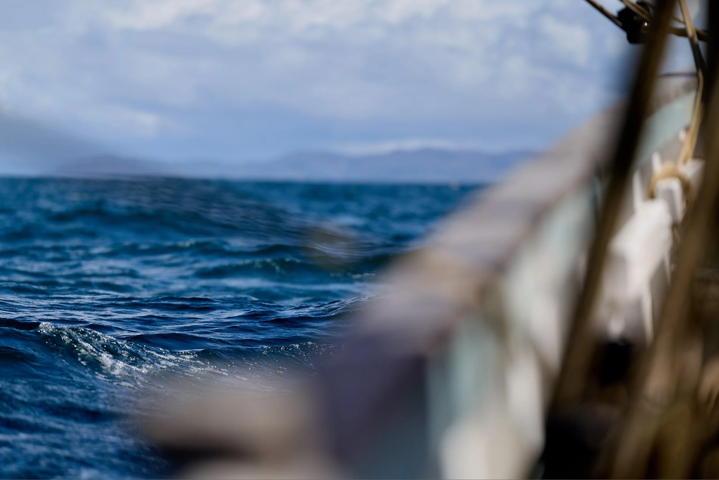 View of the ocean with blue water and distant landmass under cloudy sky, taken from a boat or ship.