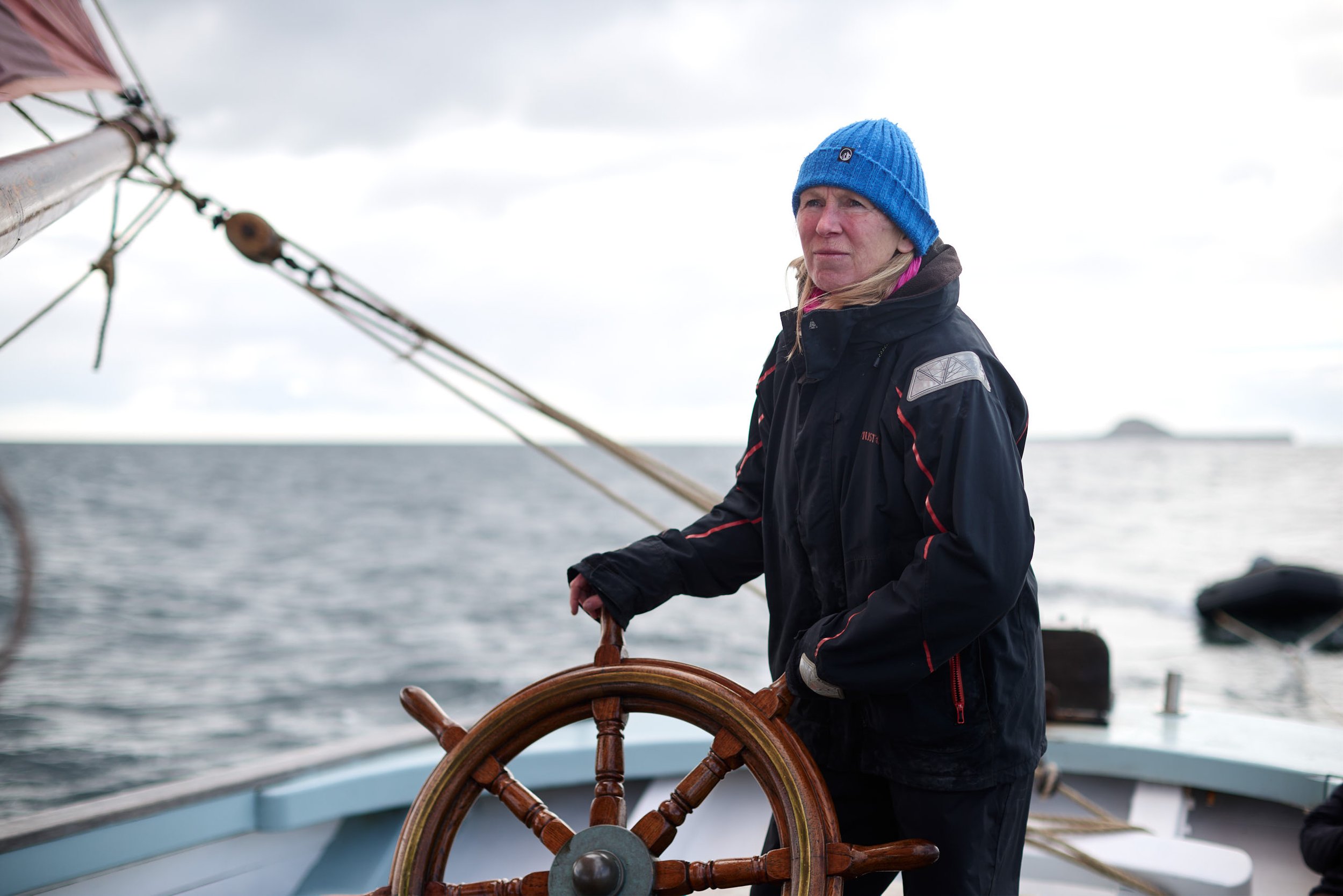 A person wearing a blue beanie and black jacket at the helm of a sailboat, steering with a wooden ship's wheel on a cloudy day at sea.