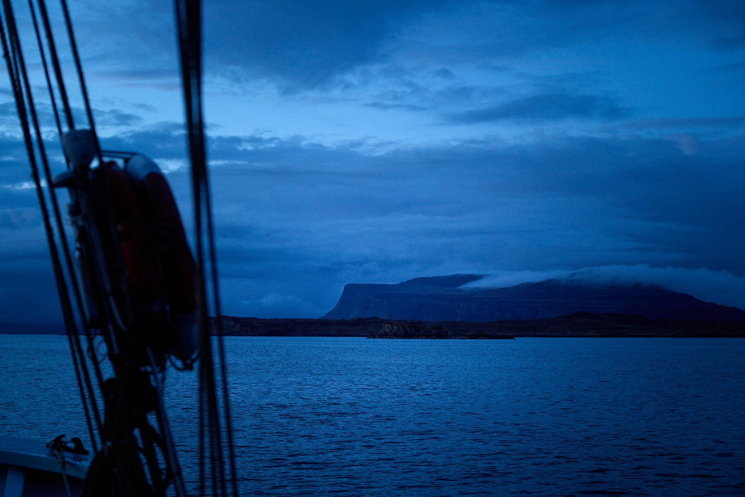 View of a calm body of water with distant cliffs and a cloudy sky, seen from the deck of a boat with parts of the boat's structure visible in the foreground.