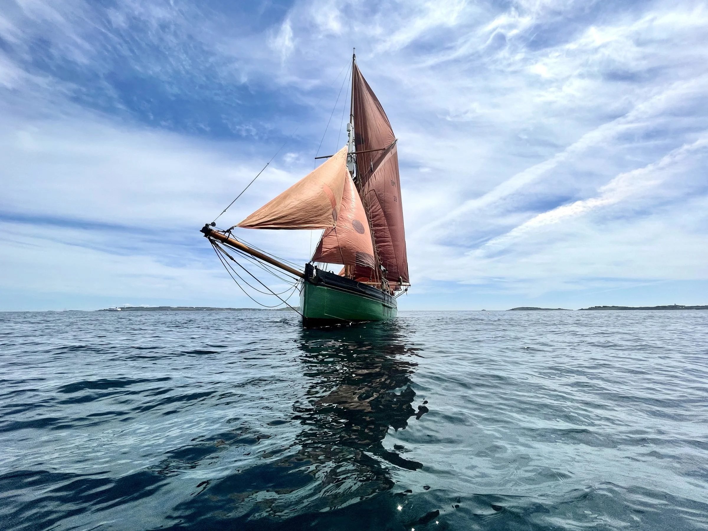 A sailing ship with brown and green sails floating on calm water under a partly cloudy sky.