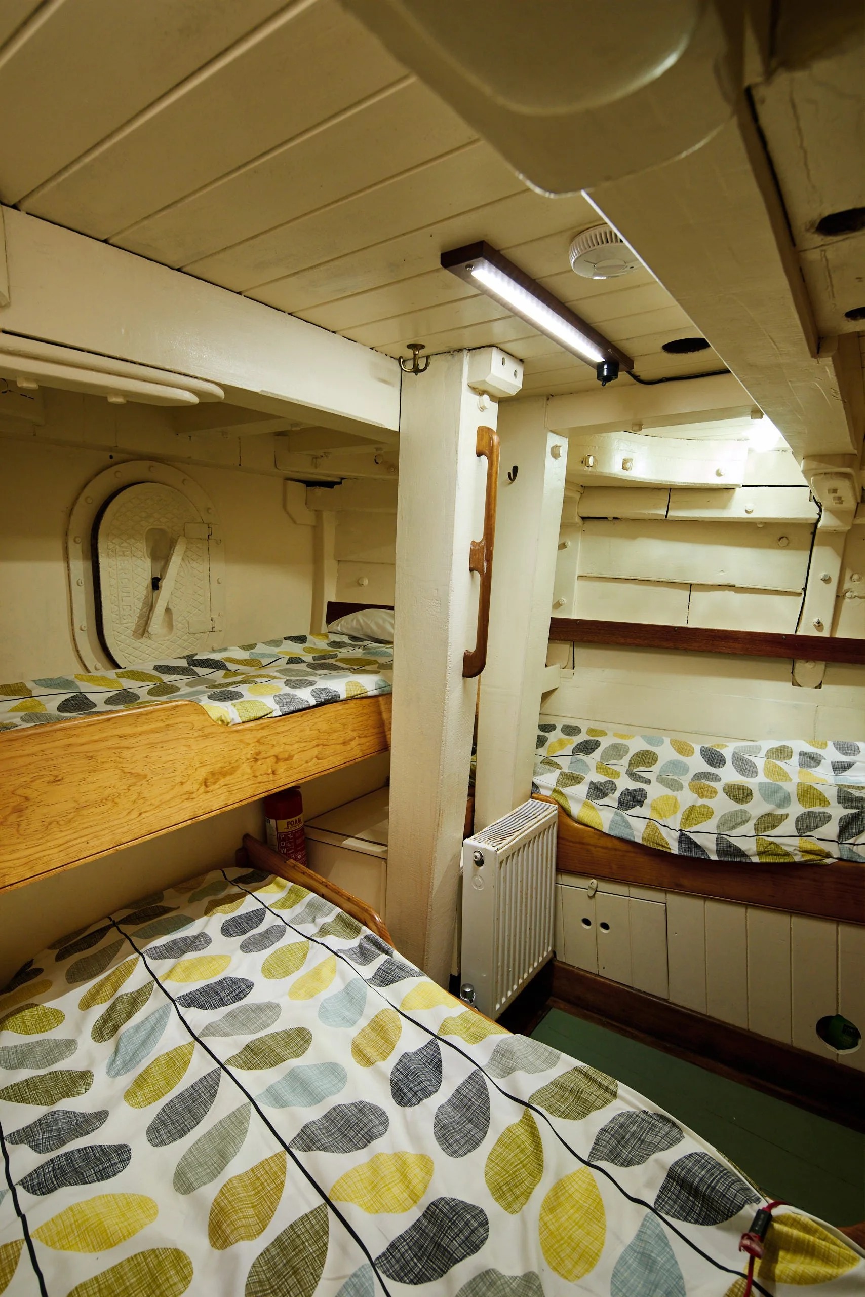 Multiple beds with patterned bedspreads in a cozy, wooden bunk room with a wood ceiling, hooks, lamp, and radiator.