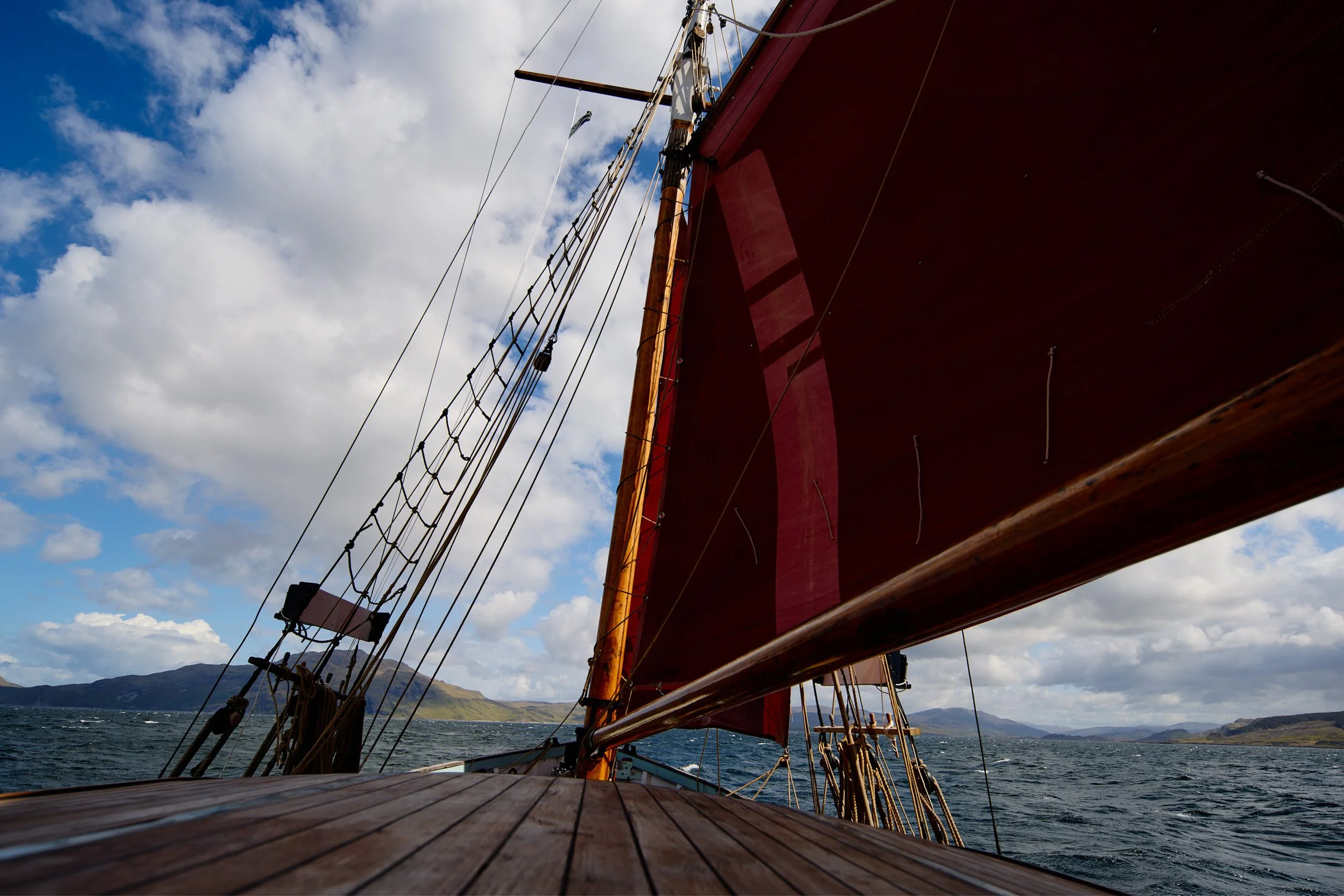 A view from the deck of a boat with a red sail, looking up at the rigging against a blue sky with clouds and distant mountains on the horizon.