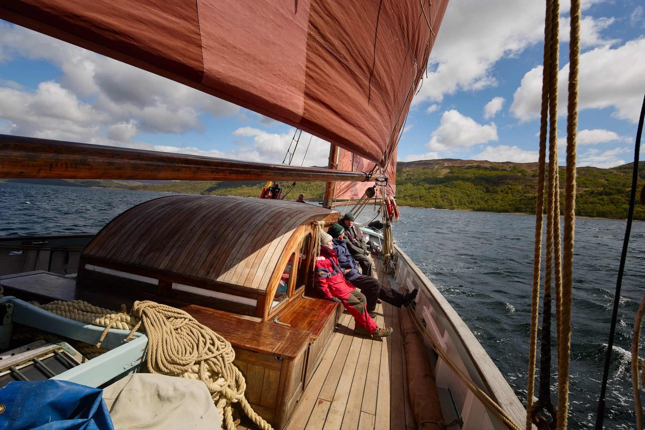 People sitting on the deck of a wooden sailing boat with large red sails, navigating through water with green hills in the background on a partly cloudy day.