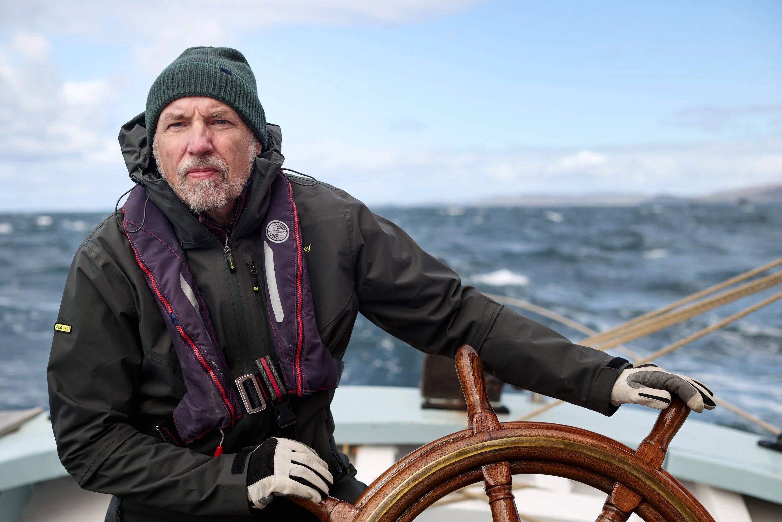 An older man with a gray beard and wearing a green knit hat and a black waterproof jacket, steering a boat by holding the wooden steering wheel. The background shows choppy ocean waters and a cloudy sky.