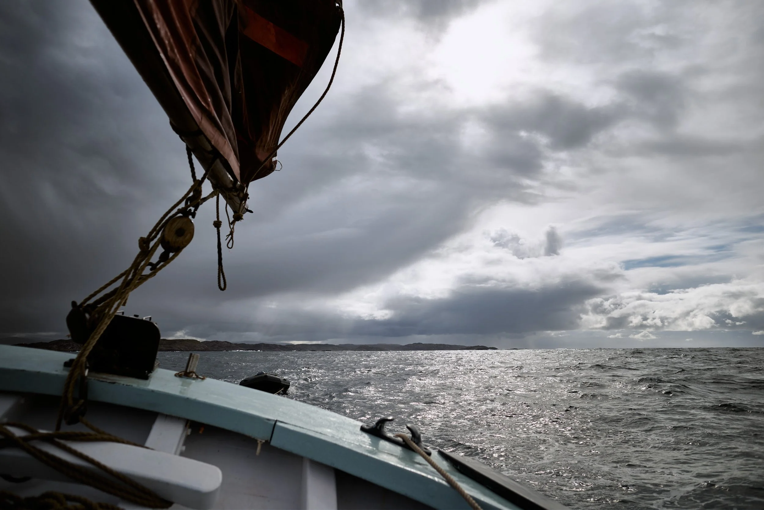View from a boat showing the bow, with ropes and part of the side, on choppy water under a cloudy sky, with distant land on the horizon.
