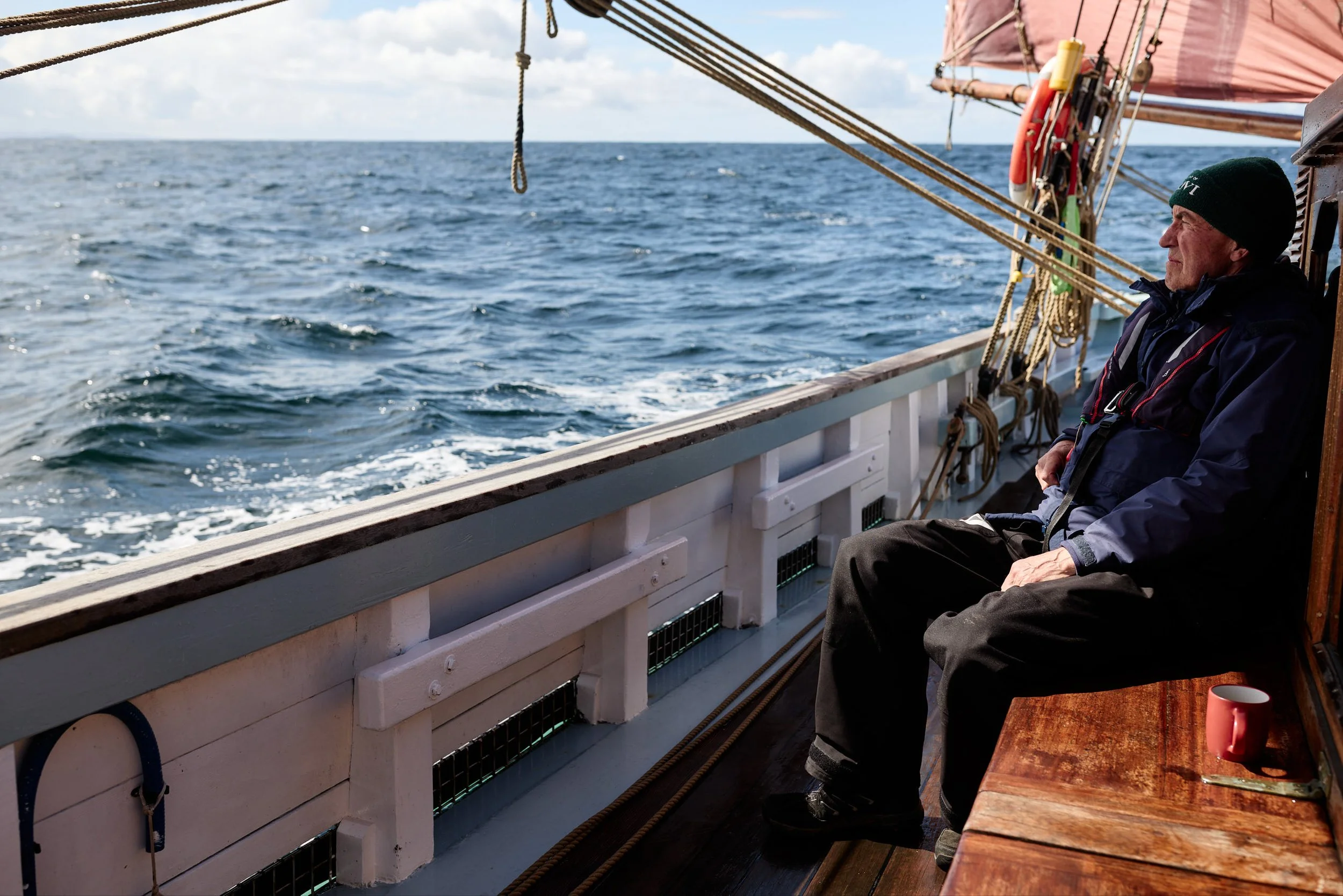 An older man wearing a black beanie and outdoor jacket sitting on a boat deck, looking out at the ocean, with ropes and boat equipment visible above and beside him.
