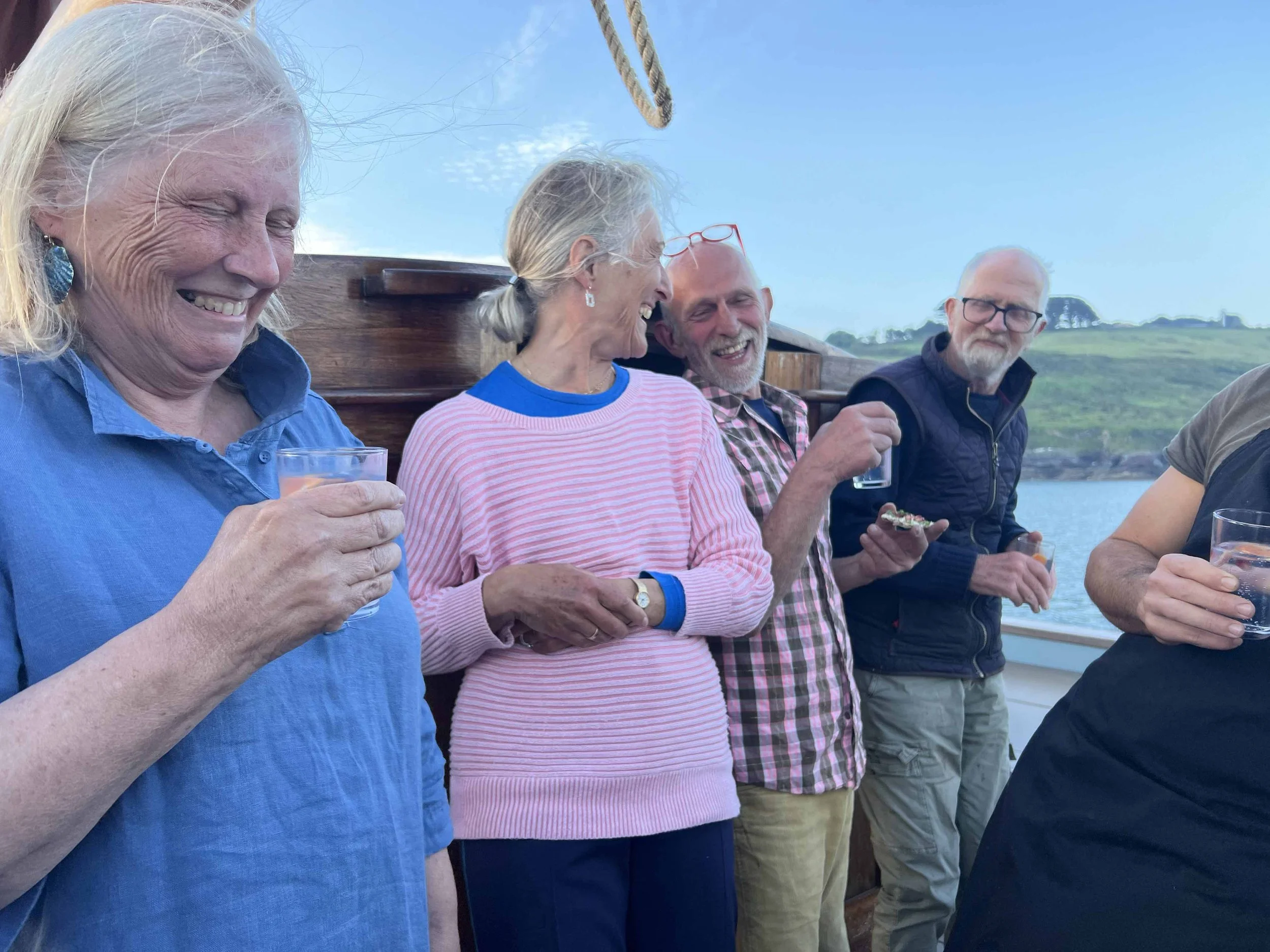 A group of elderly people standing outdoors near water, smiling and holding drinks, enjoying a social gathering on a sunny day with a blue sky and green landscape in the background.