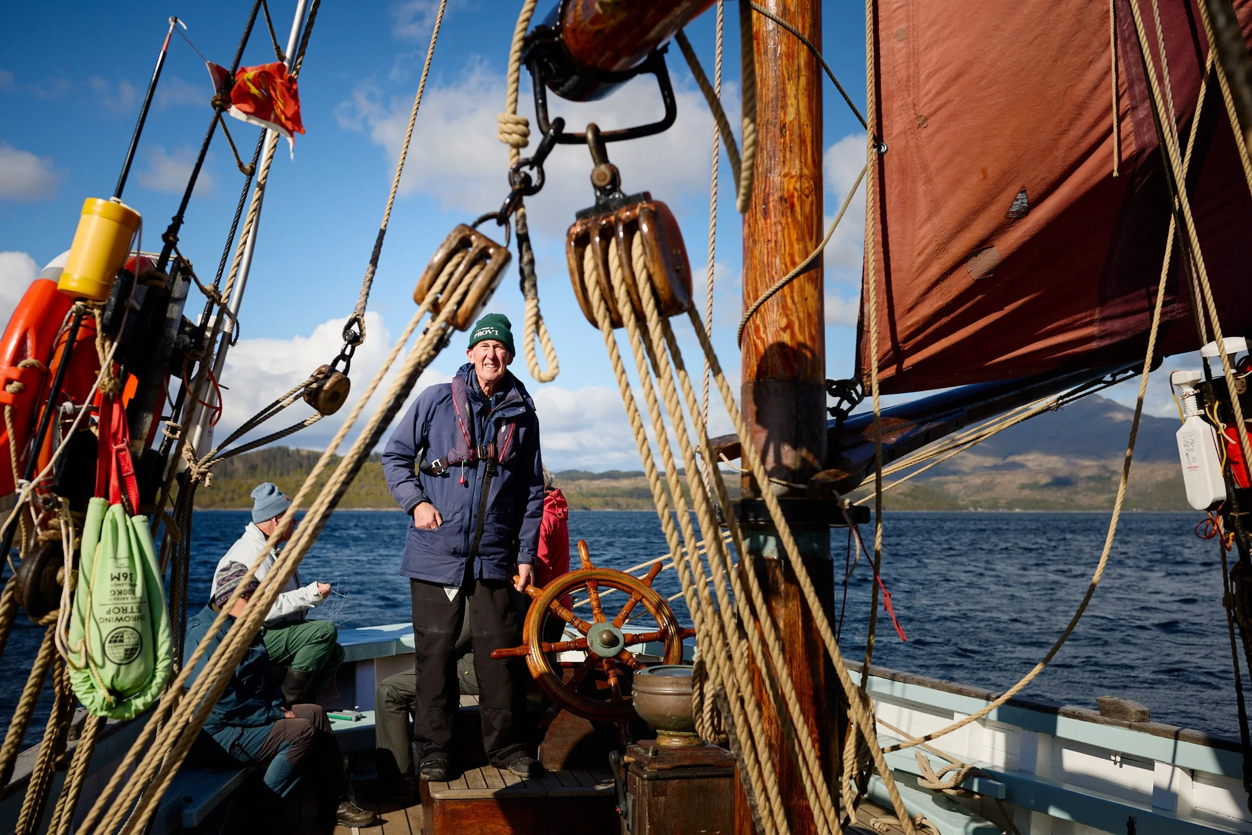 A group of people sailing on a boat with wooden steering wheel and rigging, with a scenic water and mountain background under a partly cloudy sky.