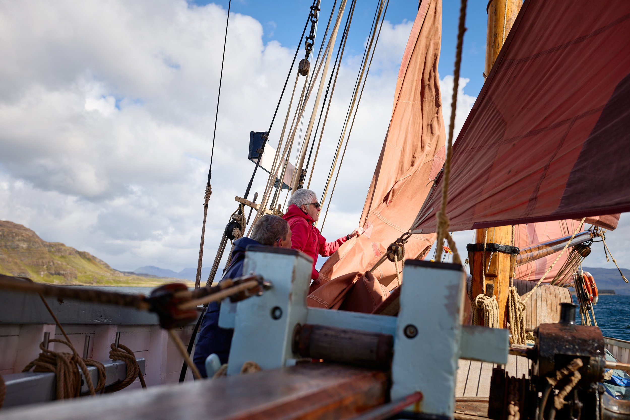 People on a sailboat adjusting sails with a scenic landscape and cloudy sky in the background.