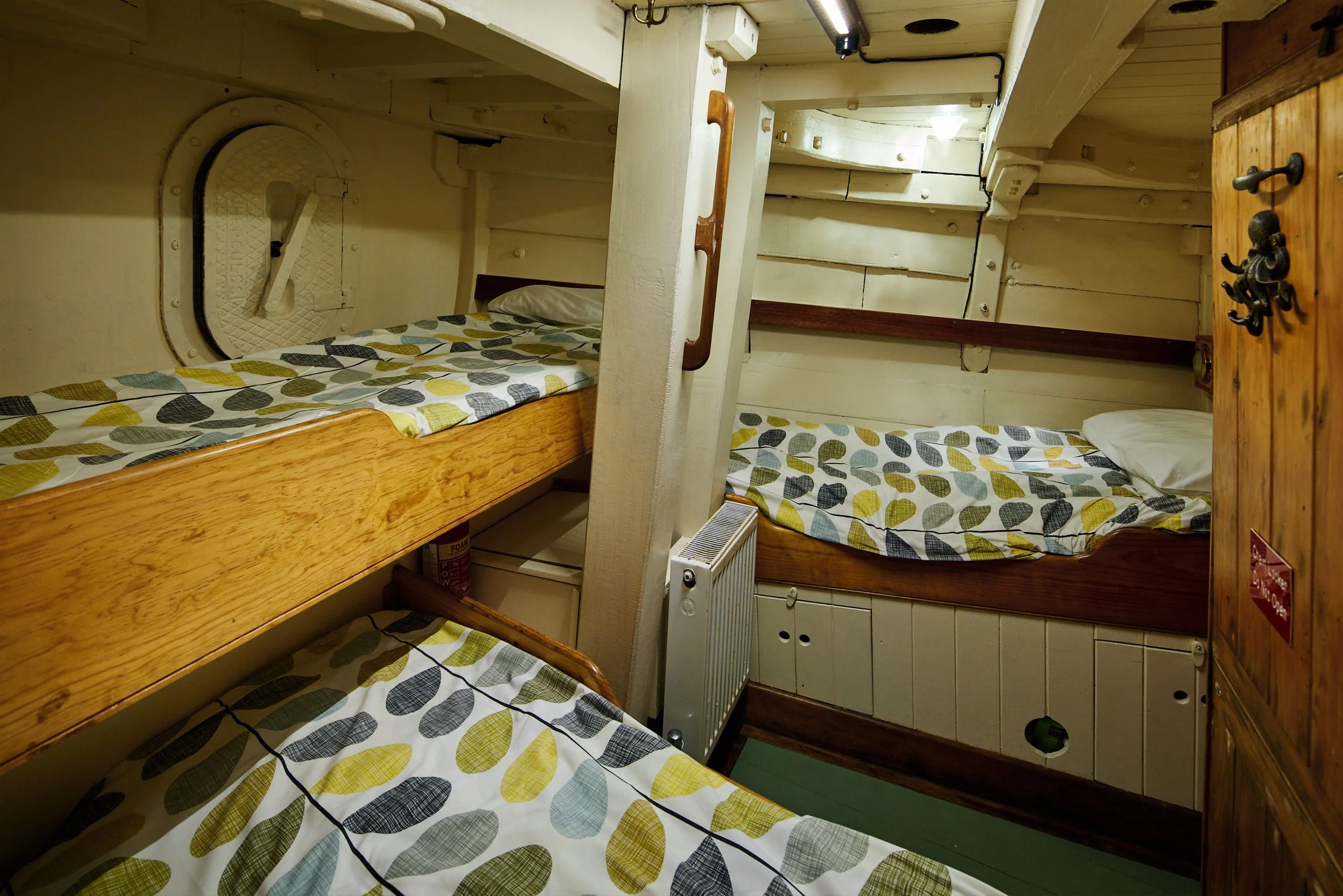 Interior of a small room with wooden beds, featuring leaf-patterned bed covers, a wooden cabinet with hooks, and a small white heater.