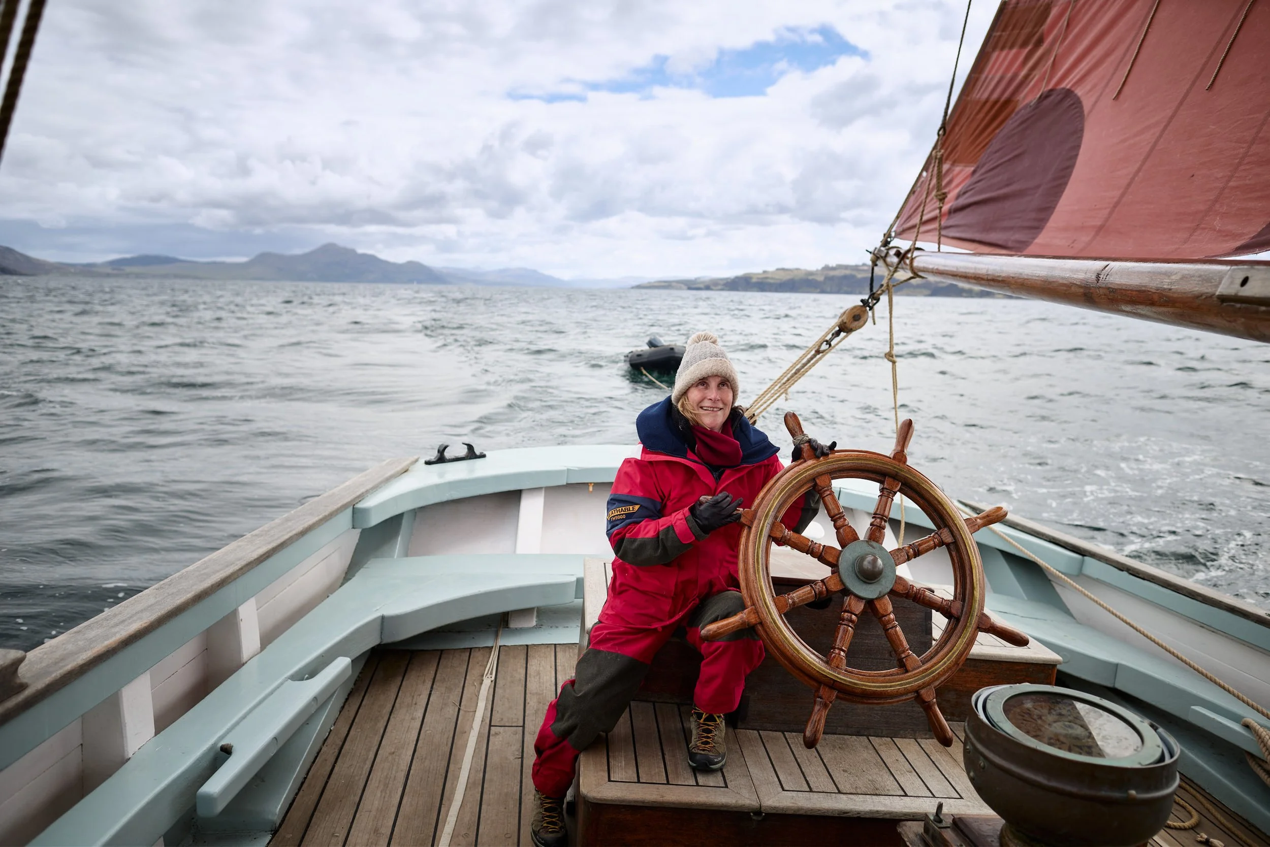 A woman with blonde hair, wearing a beige knit hat, red and black waterproof jacket, and gloves, steering a sailboat on a body of water with mountains and cloudy sky in the background.