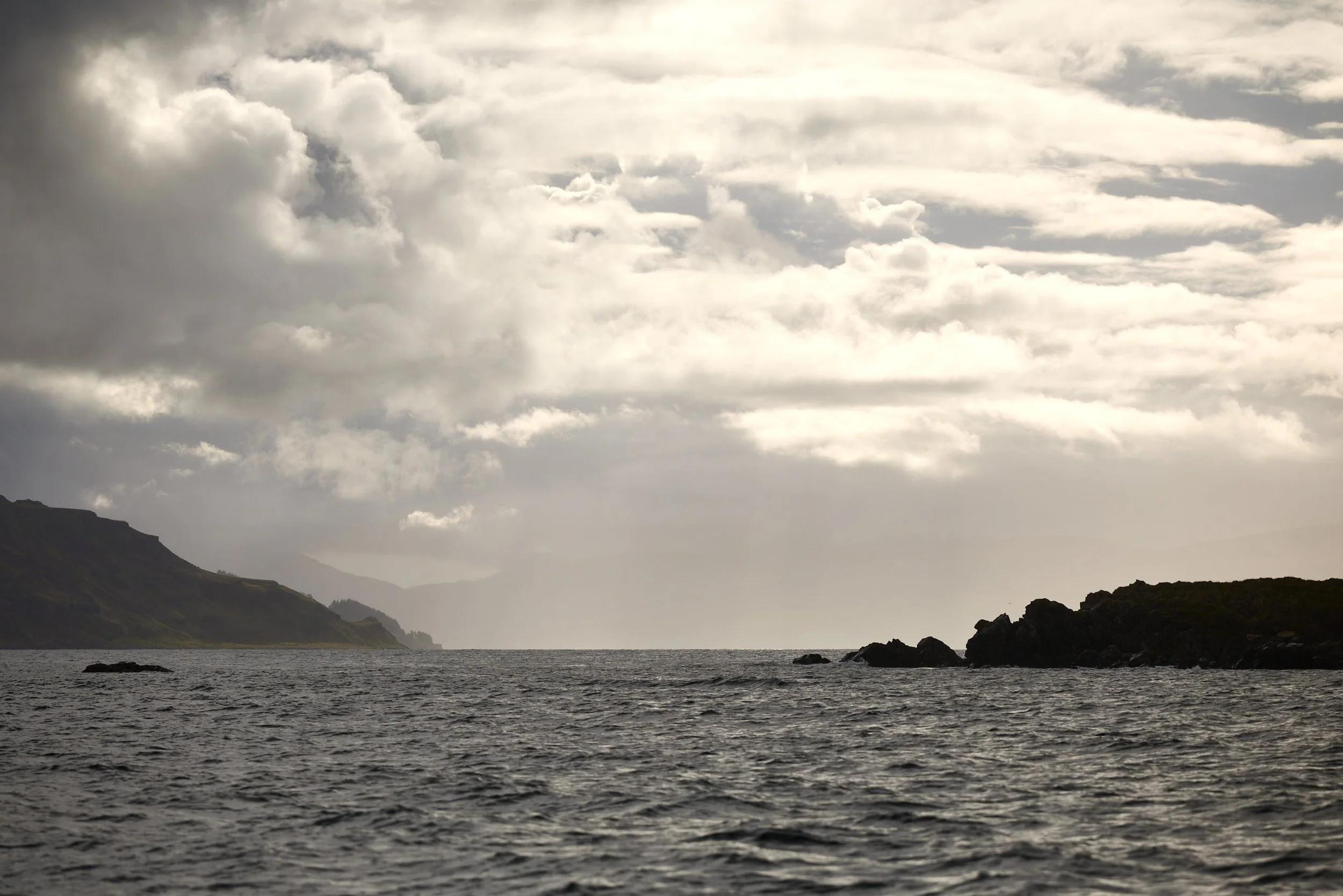A landscape scene with a body of water in the foreground, rocky coastline, and hills in the background. The sky is overcast with thick, gray clouds.