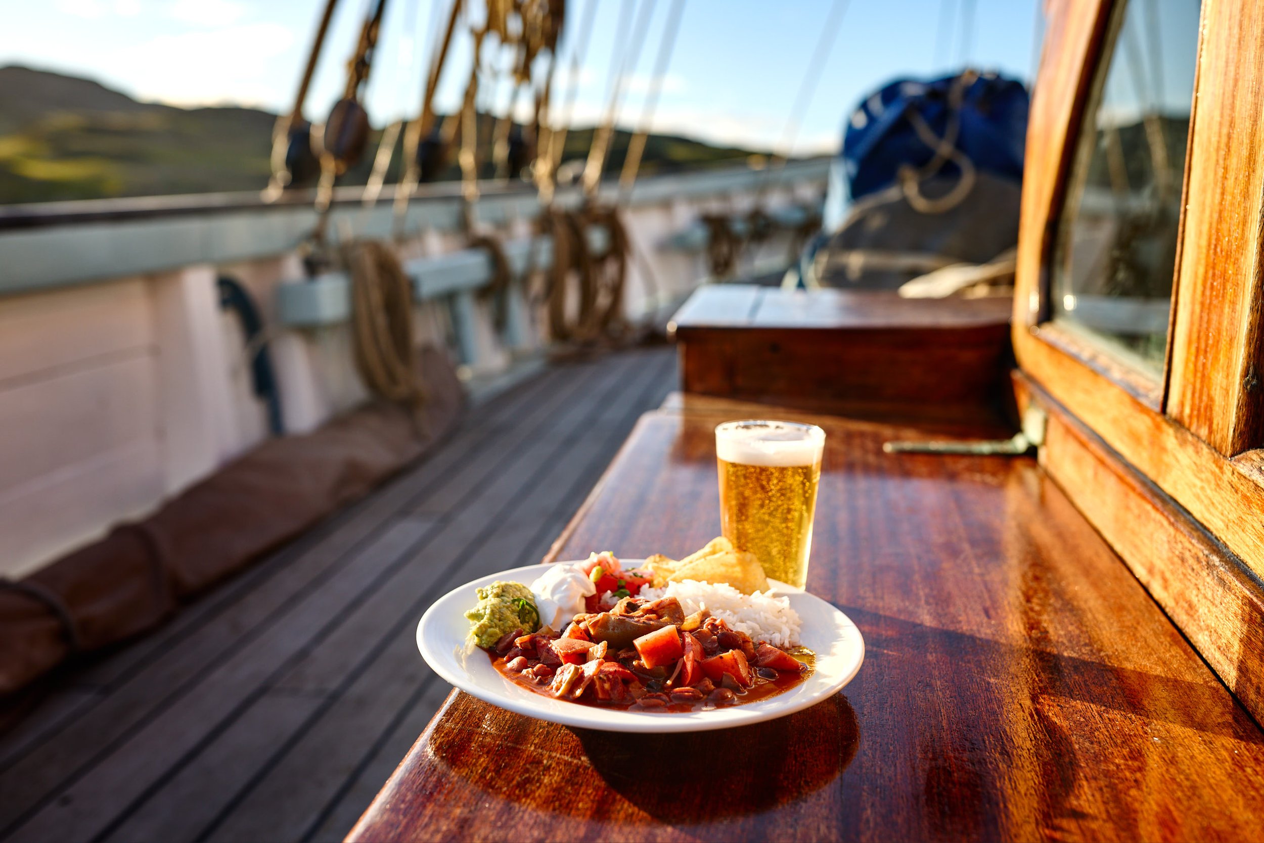 A plate of food with rice, vegetables, and meat in the foreground on a wooden table, a glass of beer, and a boat deck with ropes and a blue bag in the background during sunset.