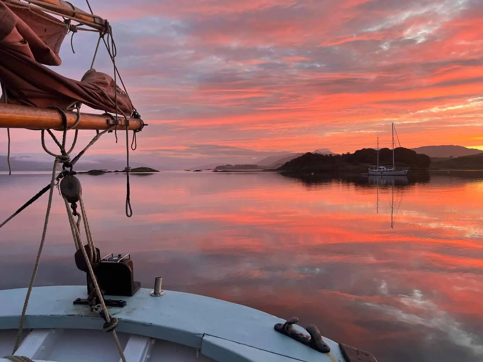View from Provident at sunset showing calm water, two sailboats in the distance, an island, and a colorful sky with pink and orange clouds.