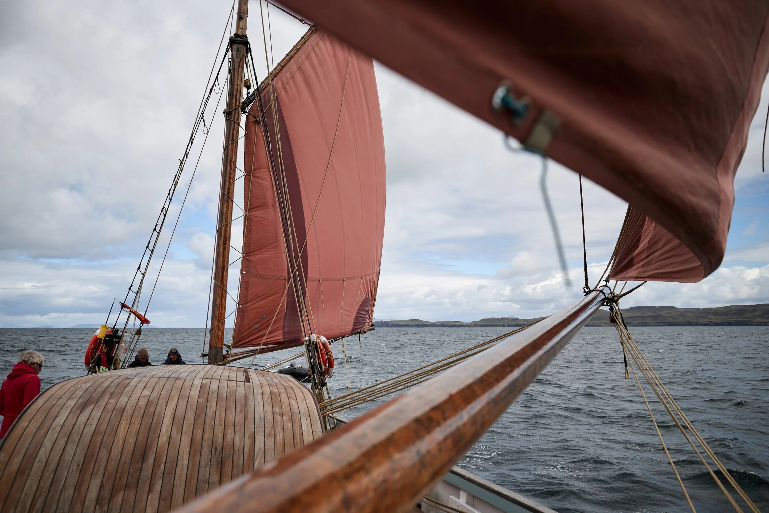 View from a sailboat with reddish sails on a cloudy day, showing people on the deck and land in the distance.
