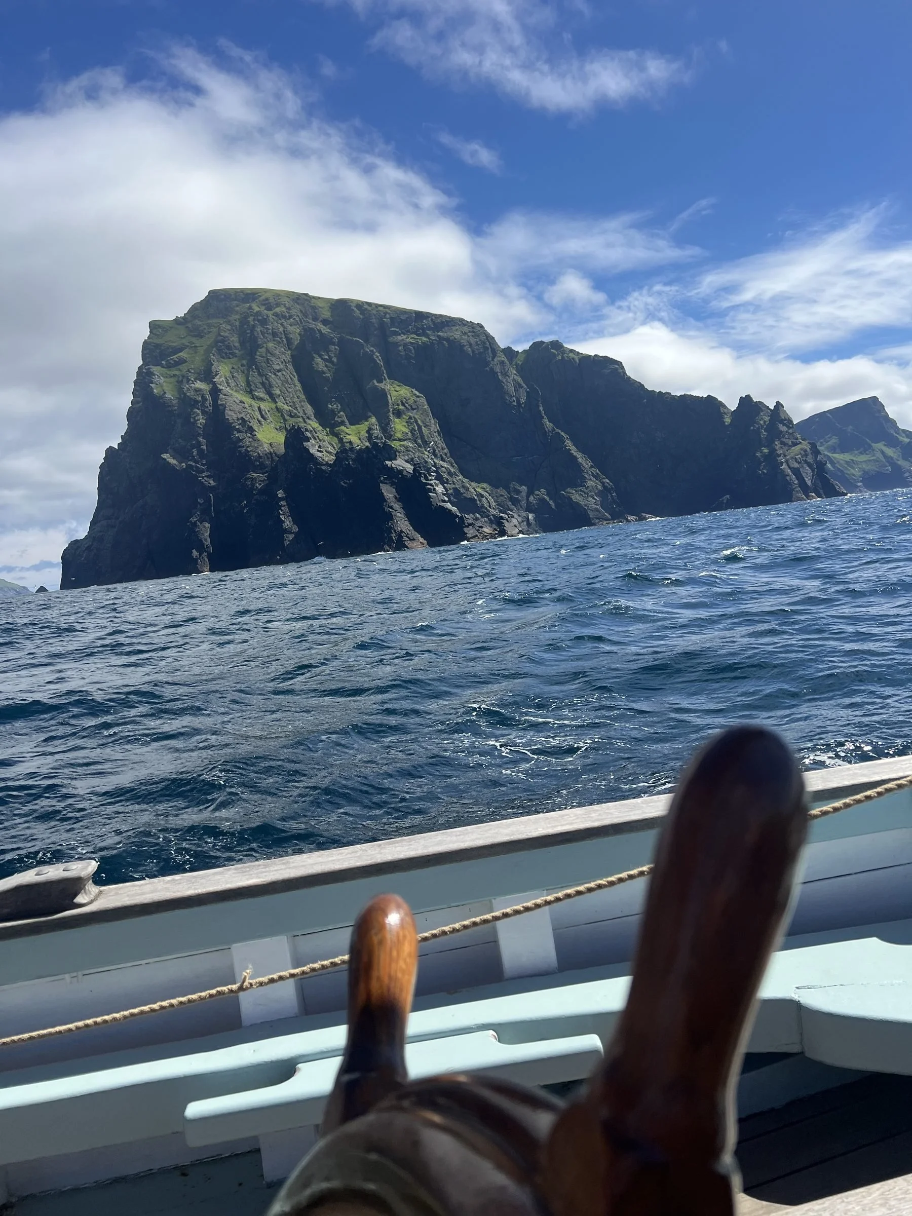 View of St Kilda with green patches, seen from a boat on the water under a partly cloudy sky.
