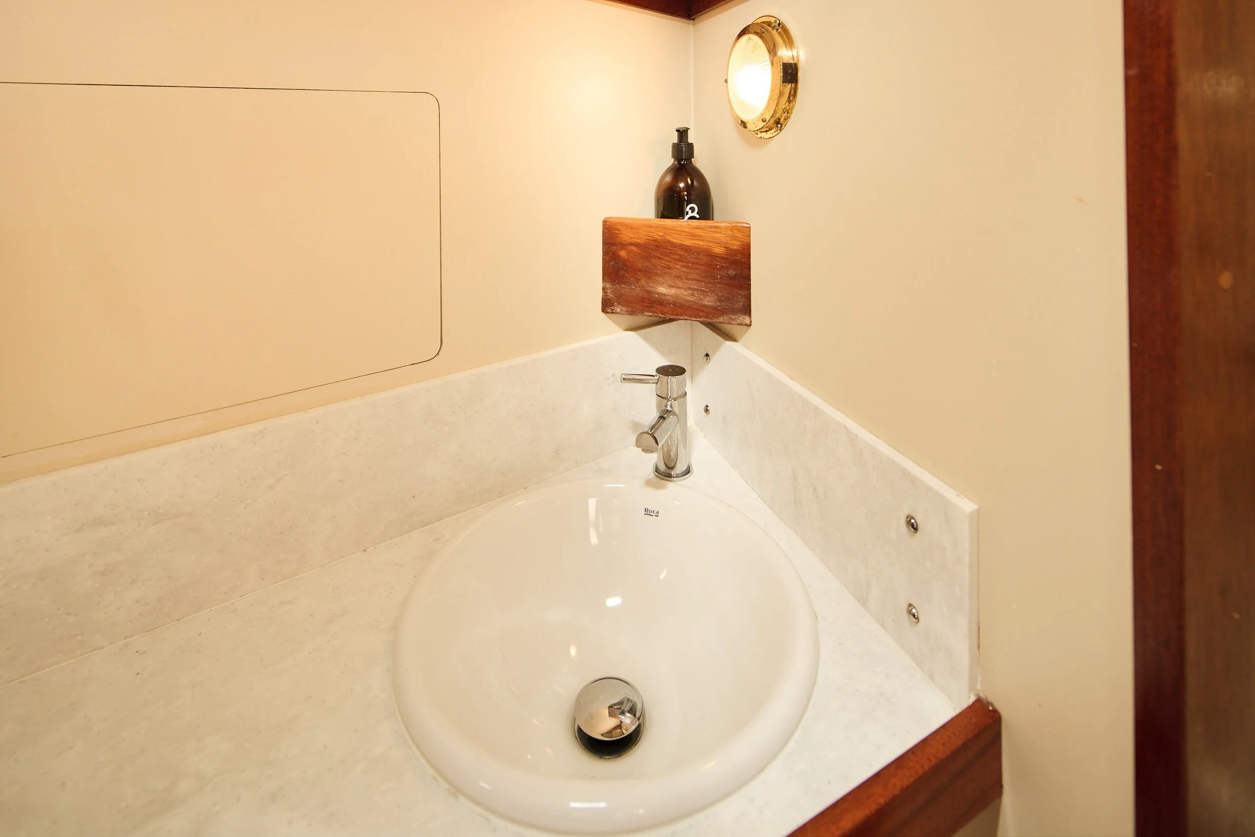 Bathroom sink with a beige countertop, a silver faucet, a round silver drain, and a small shelf above holding a dark brown soap dispenser. A gold-framed round mirror is mounted on the wall above the shelf. Part of a wooden doorframe is visible on the right.