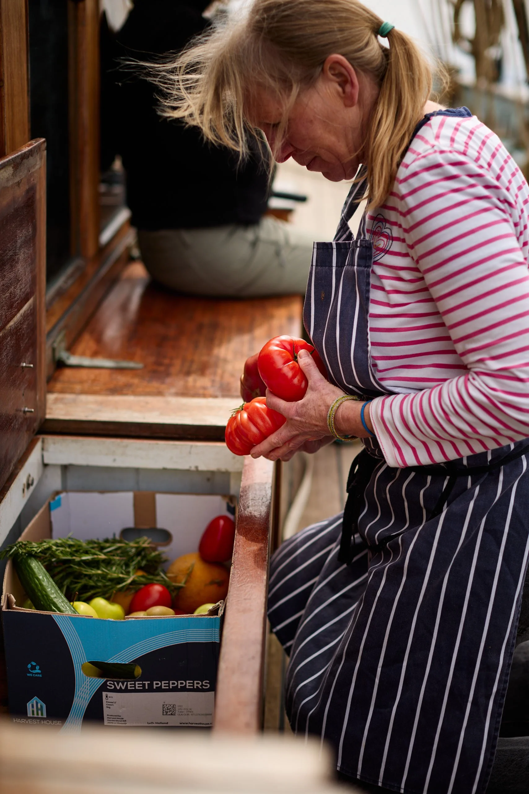 A woman wearing a striped shirt and apron holding a red tomato in a produce stand with a box of various vegetables and fruits.