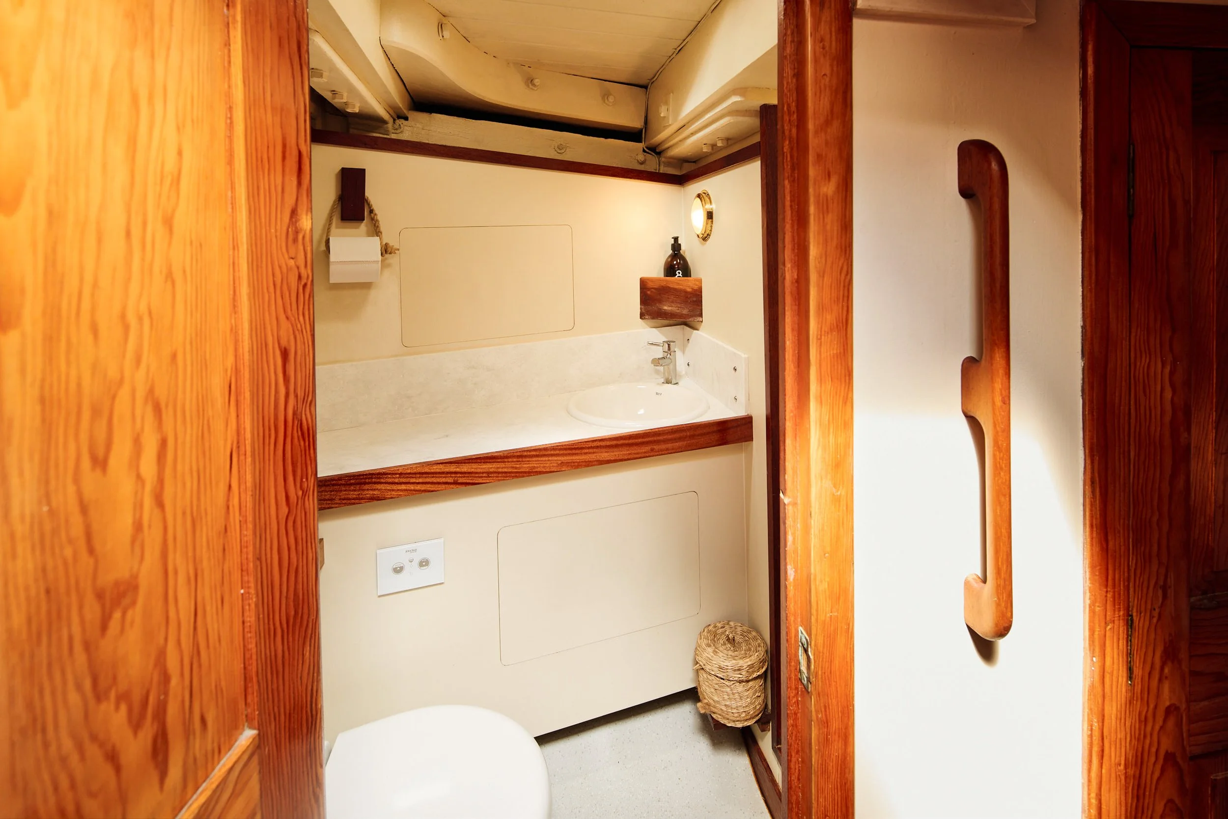 Small bathroom with a white sink, wooden accents, and a wall mirror. There is a bottle of soap, a small wooden shelf, a woven basket, and a wall-mounted paper towel holder.