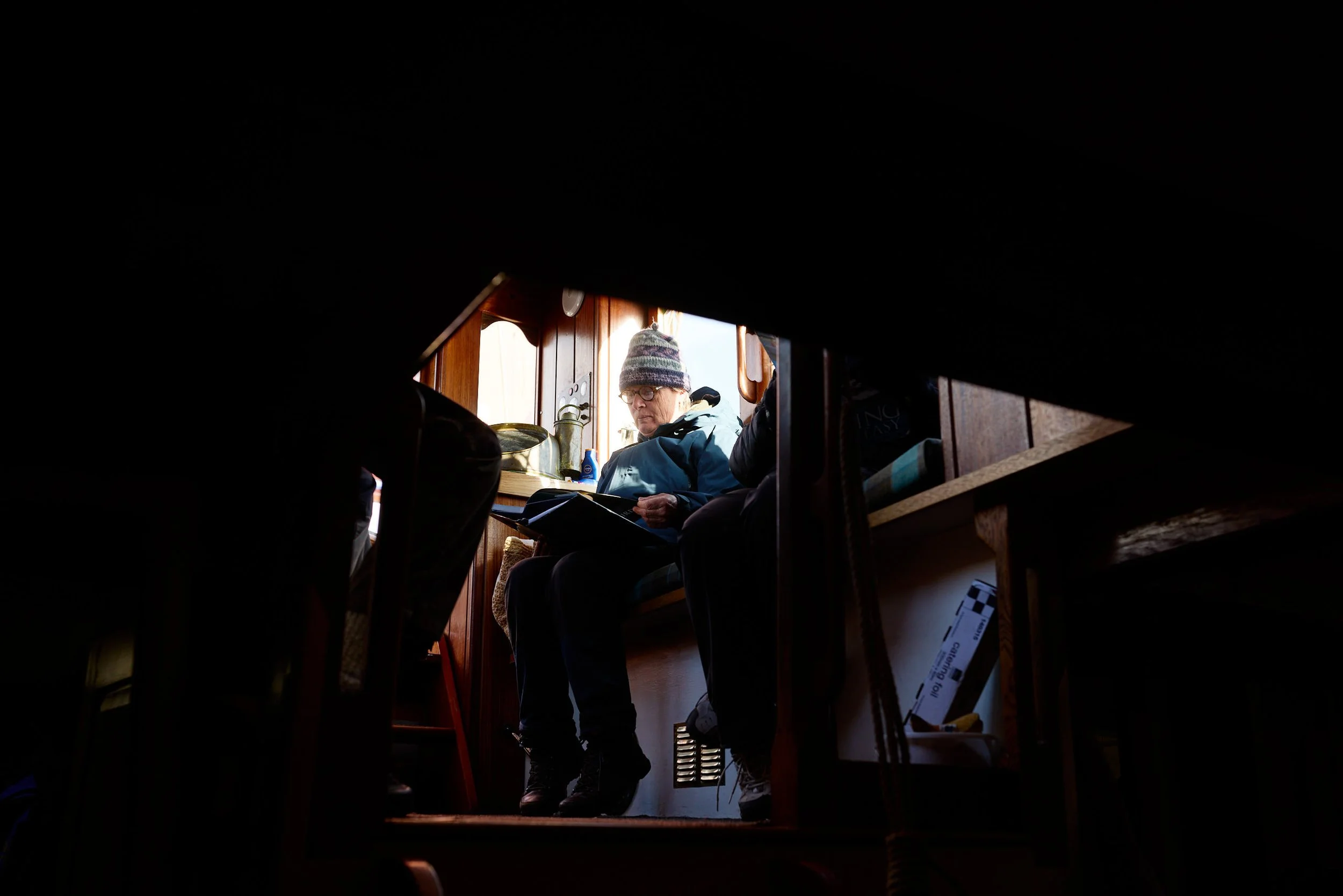 Person wearing glasses and a knit hat sitting inside a boat or cabin, looking towards the camera, with a wooden interior and natural light.